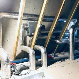 Sanerga - Montaż instalacji wentylacyjnej w poddaszu: srebrne, izolowane rury prowadzone między drewnianymi krokwiami, widoczny fragment centrali wentylacyjnej i niebieski przewód elastyczny.
