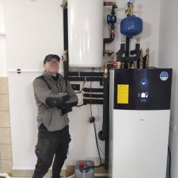 Sanerga - Uśmiechnięty instalator w okularach, z założonymi rękami, prezentuje zamontowaną pompę ciepła Daikin wraz z zasobnikiem CWU i osprzętem w kotłowni z beżowymi płytkami podłogowymi.