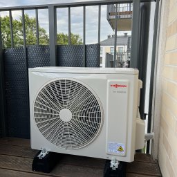 Sanerga - Biała jednostka zewnętrzna klimatyzatora Viessmann DC Inverter, zamontowana na czarnych podkładkach na drewnianym balkonie z szarą balustradą, widok na budynek mieszkalny w tle.