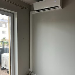 Sanerga - Biały klimatyzator marki Viessmann zamontowany na szarej ścianie w pokoju z balkonem; widoczna jednostka zewnętrzna na balkonie i zabezpieczona podłoga.