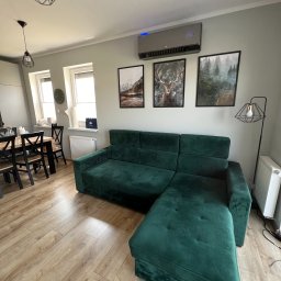 Sanerga - Szary klimatyzator ścienny z niebieskim podświetleniem, zamontowany nad zieloną, welurową sofą w nowoczesnym salonie z drewnianą podłogą i geometrycznymi lampami.