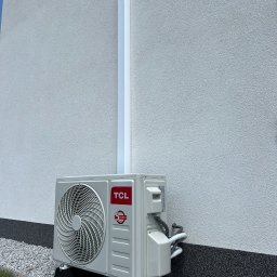 Sanerga - Zewnętrzna jednostka klimatyzacji, model TCL, zamontowana przy ścianie budynku z szarym tynkiem, na podłożu z drobnych kamieni. Widoczna biała rura odprowadzająca skropliny.