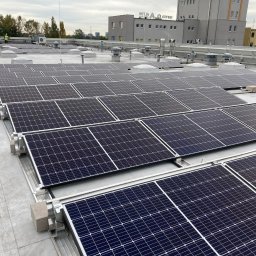 Sanerga - Rząd paneli słonecznych na dachu budynku przemysłowego, widoczny pracownik w odblaskowej kamizelce, szare niebo.