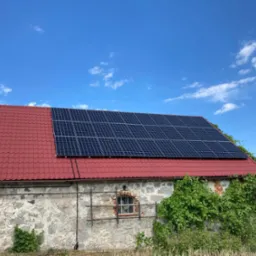 Panele słoneczne na czerwonym dachu starego, kamiennego budynku gospodarczego w słoneczny dzień.