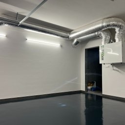 Sanerga - Biała jednostka rekuperacyjna WANAS 615 Hi-Tech z aluminiowymi przewodami wentylacyjnymi na suficie w jasnym pomieszczeniu z ciemną podłogą.