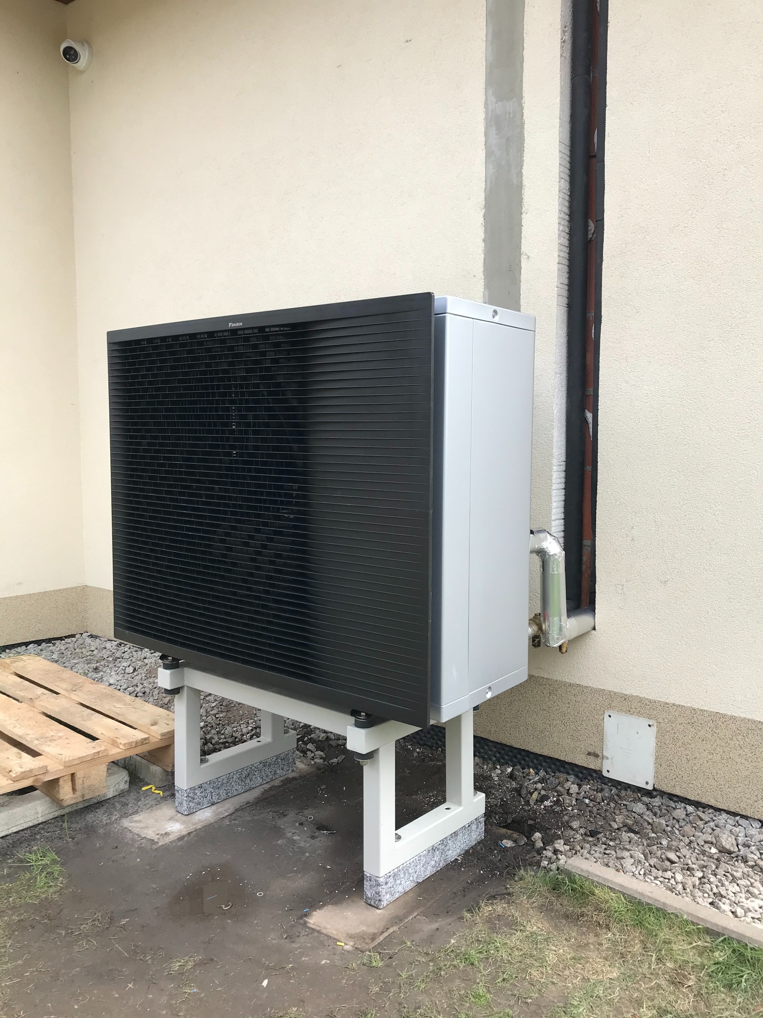 Zewnętrzna jednostka pompy ciepła Daikin, zamontowana na stalowej konstrukcji z granitowymi podkładkami, przy ścianie budynku z widocznymi rurami instalacji i kamerą monitoringu.