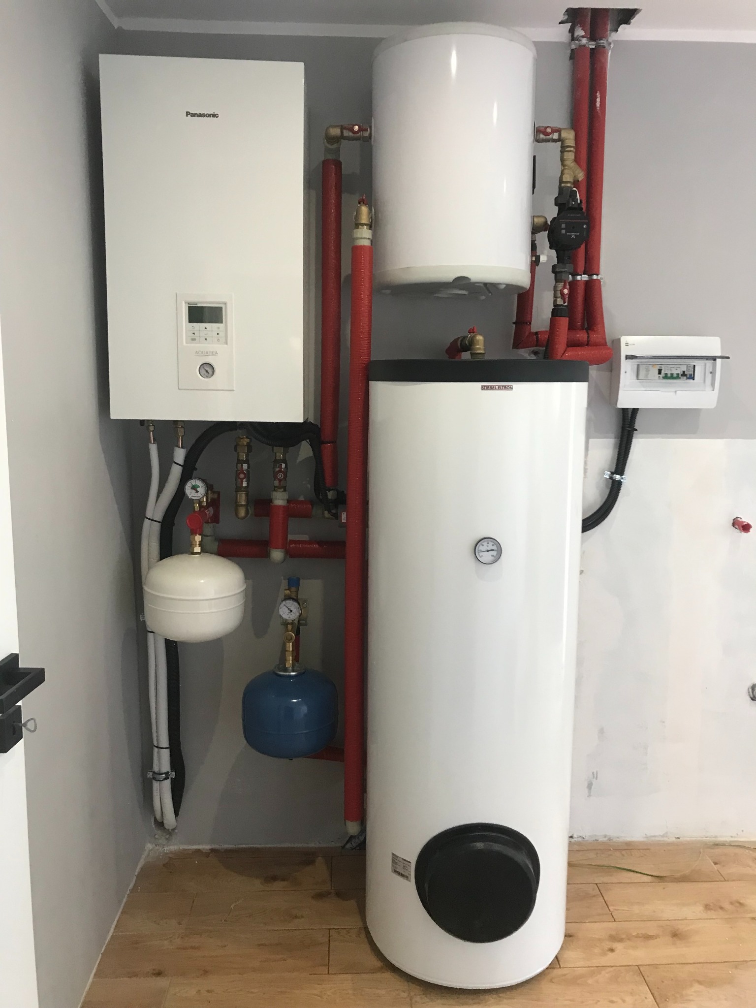 Biały bojler Stiebel Eltron z podłączoną instalacją hydrauliczną, obok biała pompa ciepła Panasonic Aquarea z widocznymi manometrami i rurami w izolacji termicznej, całość zamontowana na drewnianej...