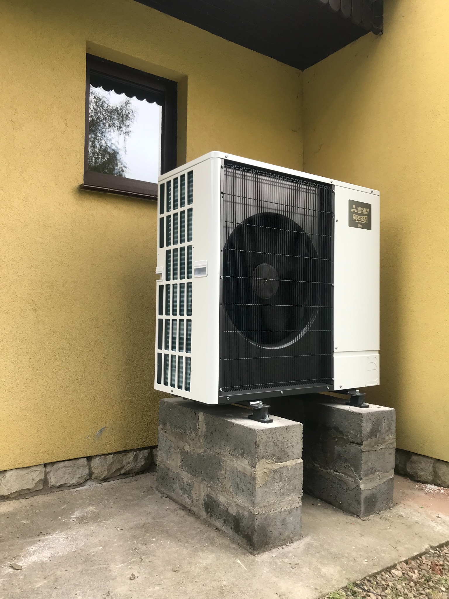 Biała jednostka zewnętrzna pompy ciepła Mitsubishi Electric Zubadan R32, zamontowana na dwóch betonowych bloczkach przy żółtej ścianie budynku z oknem.