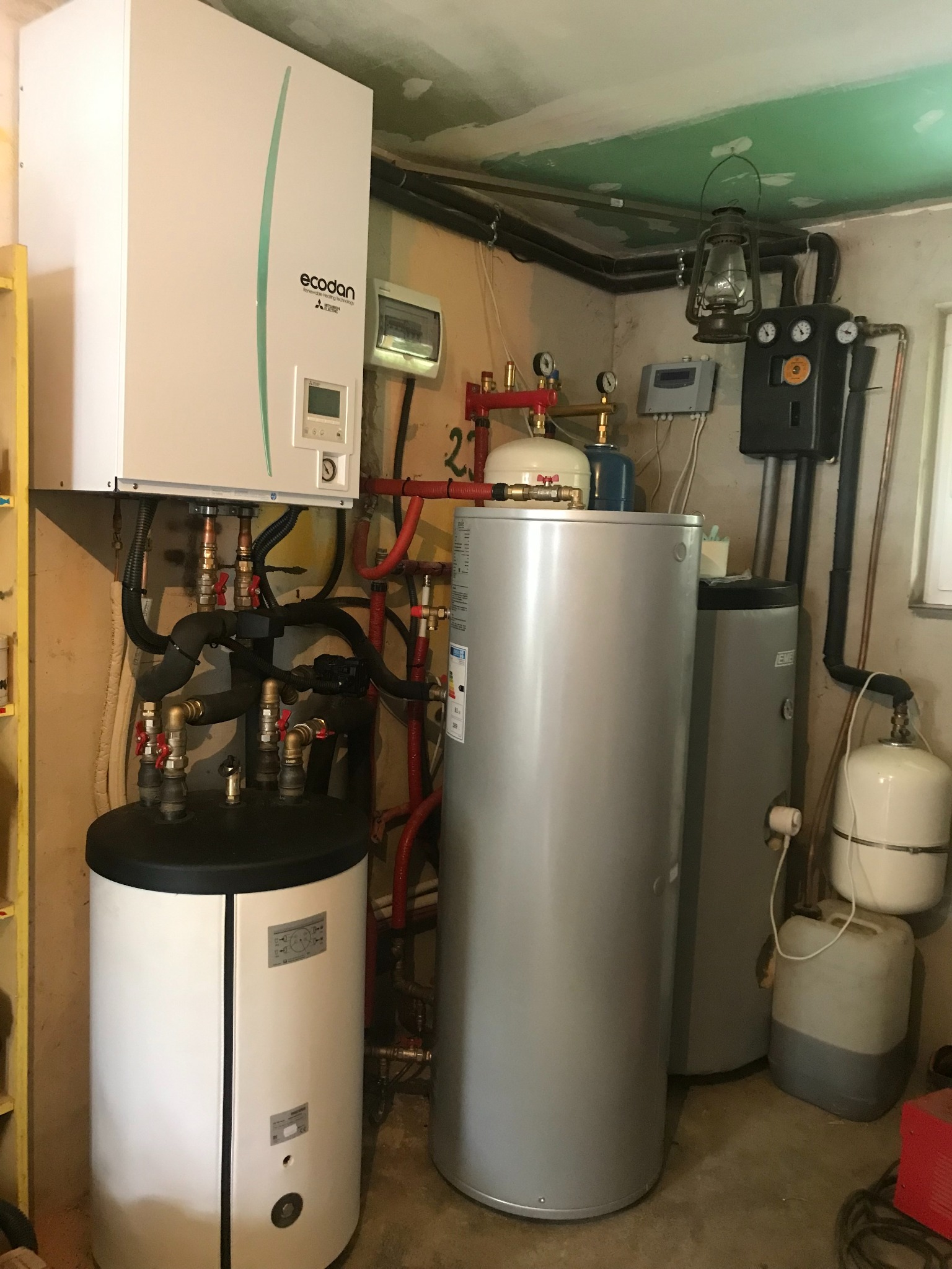 Zestaw urządzeń grzewczych w kotłowni: biała jednostka Ecodan na ścianie, obok zbiorniki akumulacyjne, widoczne rury i zawory, a także stara lampa naftowa pod sufitem.