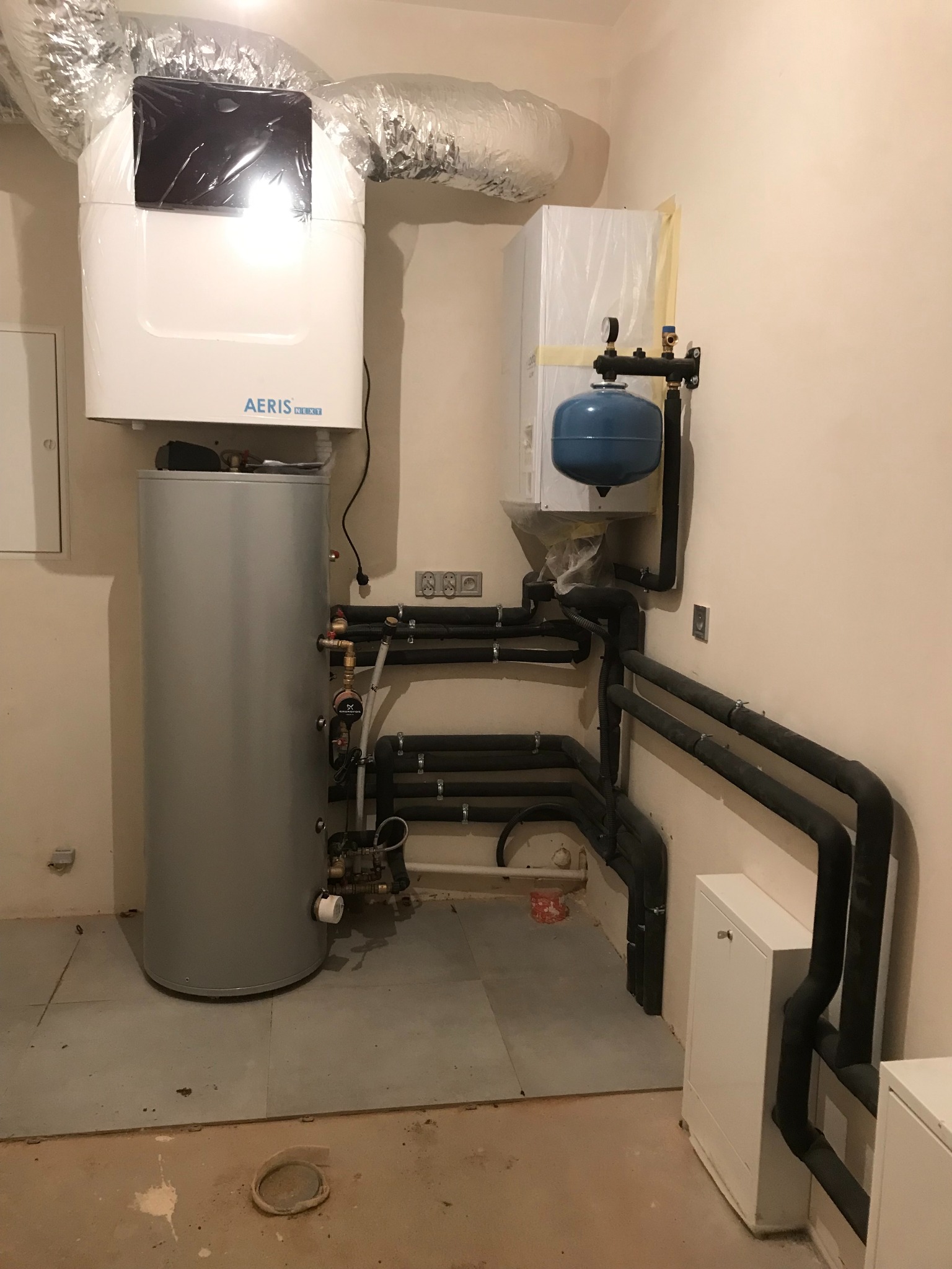 Wnętrze kotłowni z zamontowanym zbiornikiem buforowym, jednostką centralną i dodatkowym urządzeniem, połączonymi systemem rur izolowanych termicznie. Elementy częściowo w folii ochronnej.