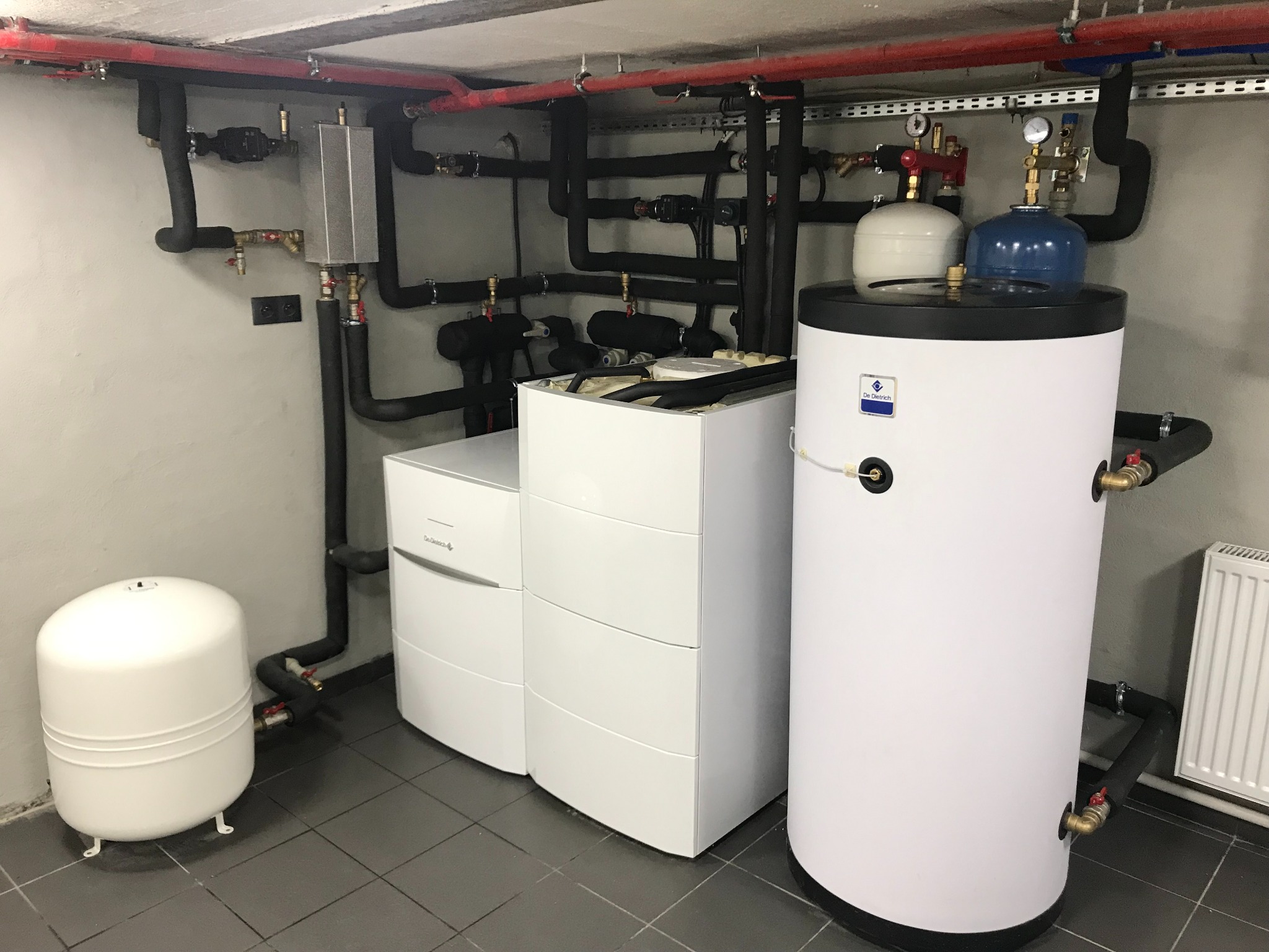Biała kotłownia z grzejnikiem, zbiornikiem buforowym i rozbudowaną instalacją rur miedzianych i czarnych, zamontowana w piwnicy na ciemnych kaflach.