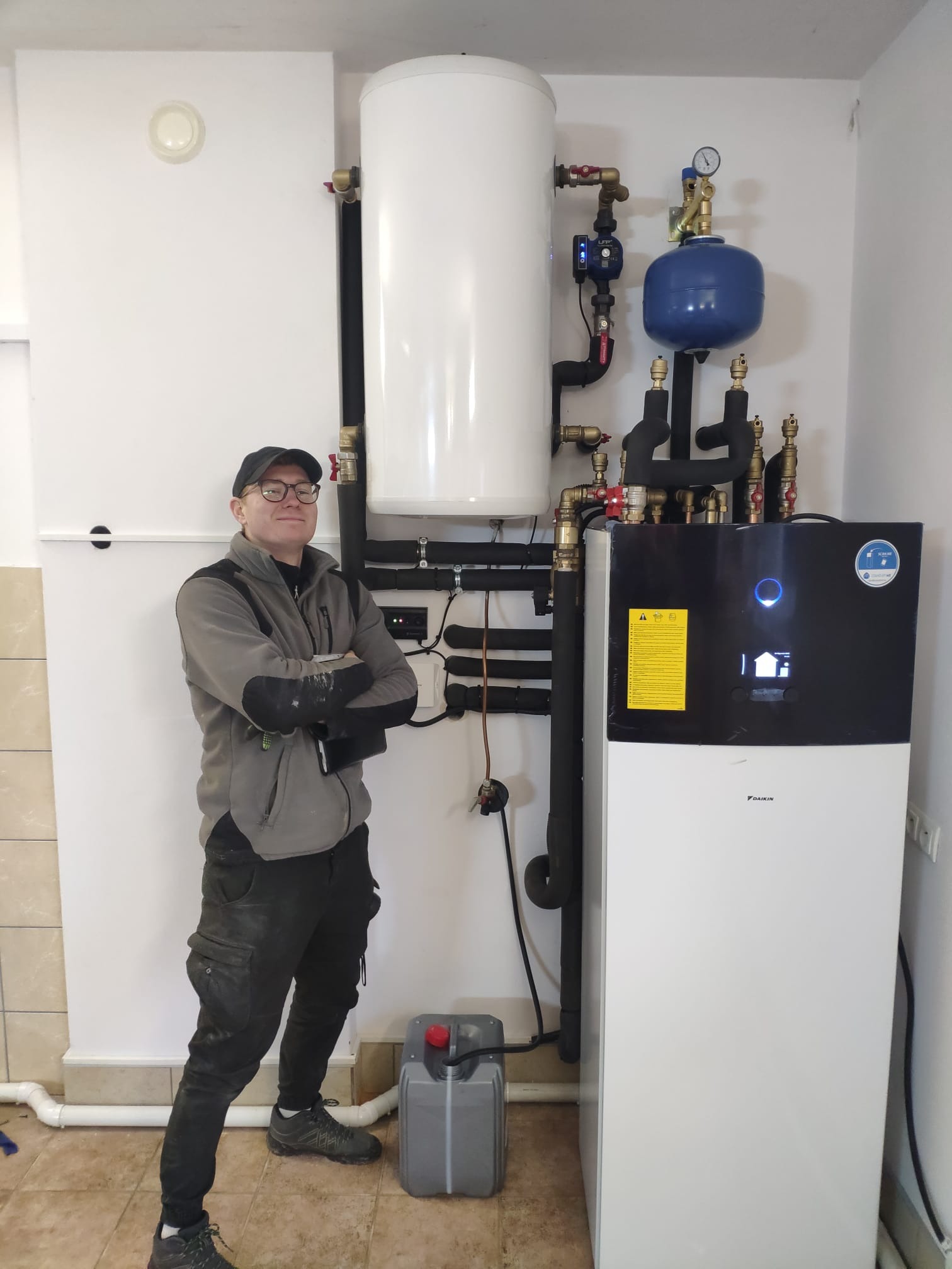 Uśmiechnięty instalator w okularach, z założonymi rękami, prezentuje zamontowaną pompę ciepła Daikin wraz z zasobnikiem CWU i osprzętem w kotłowni z beżowymi płytkami podłogowymi.