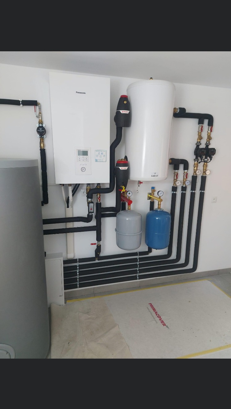 Biały wiszący piec gazowy Panasonic, zbiornik na wodę Galmet i dwa naczynia przeponowe w kolorach szarym i niebieskim połączone czarnymi rurami w kotłowni na tle białej ściany, podłoga...