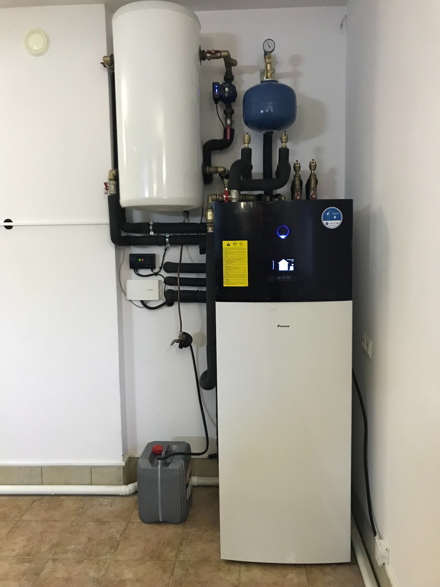 Nowoczesna, biało-czarna instalacja grzewcza Daikin z zbiornikiem, rurami i osprzętem, umieszczona w rogu pomieszczenia z beżowymi płytkami podłogowymi; obok kanister z płynem.