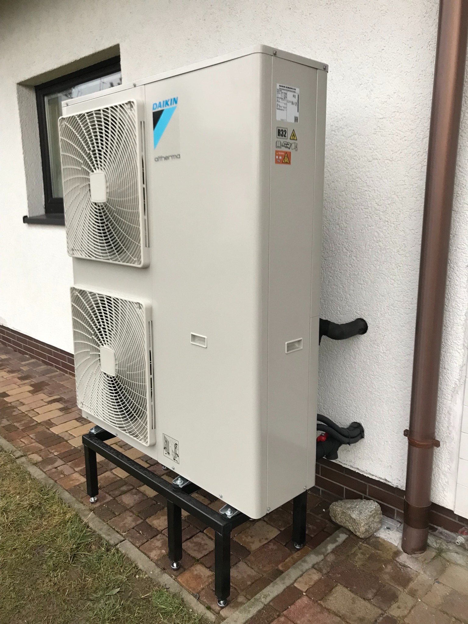 Biała jednostka zewnętrzna z dwoma wentylatorami, marki Daikin Altherma, zamontowana na czarnym, metalowym stelażu przy ścianie budynku z widoczną rurą spustową i brukowanym podłożem.
