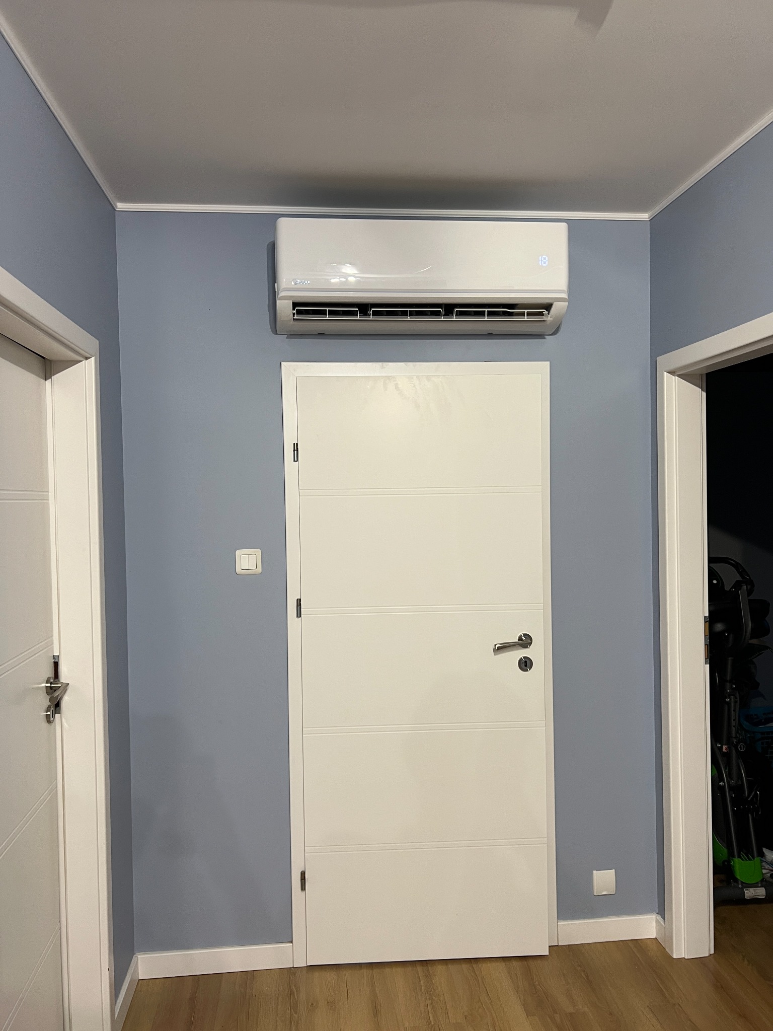 Biały klimatyzator ścienny zamontowany nad białą, panelową drzwiami w pomieszczeniu z szarą ścianą i białymi framugami.