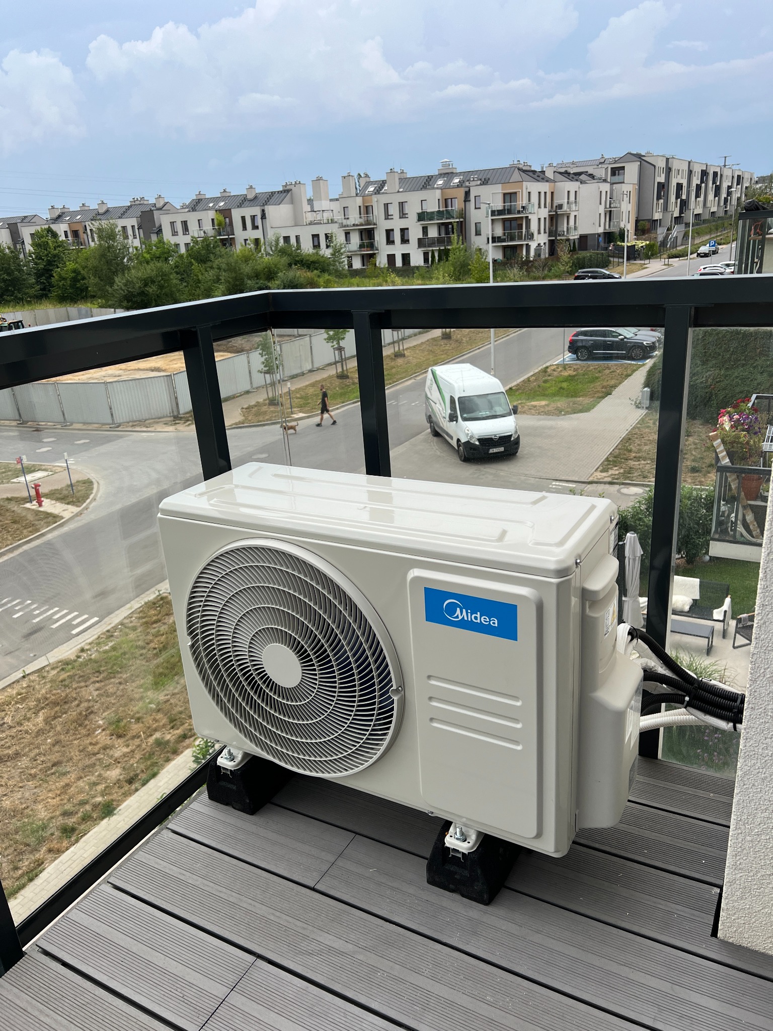Jednostka zewnętrzna klimatyzatora Midea na balkonie nowego budownictwa, widok na osiedle, ulicę i zaparkowany biały samochód dostawczy.