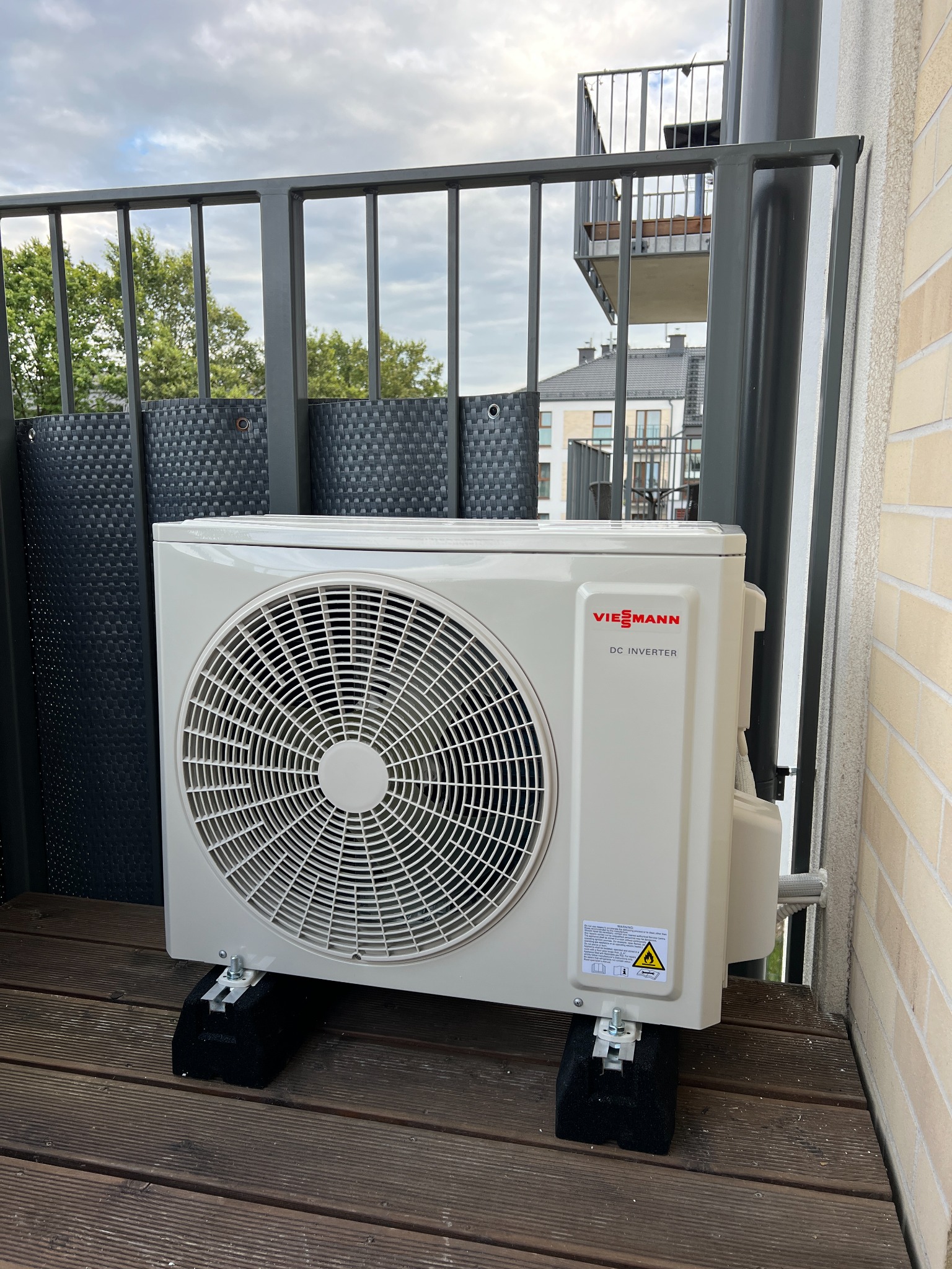 Biała jednostka zewnętrzna klimatyzatora Viessmann DC Inverter, zamontowana na czarnych podkładkach na drewnianym balkonie z szarą balustradą, widok na budynek mieszkalny w tle.