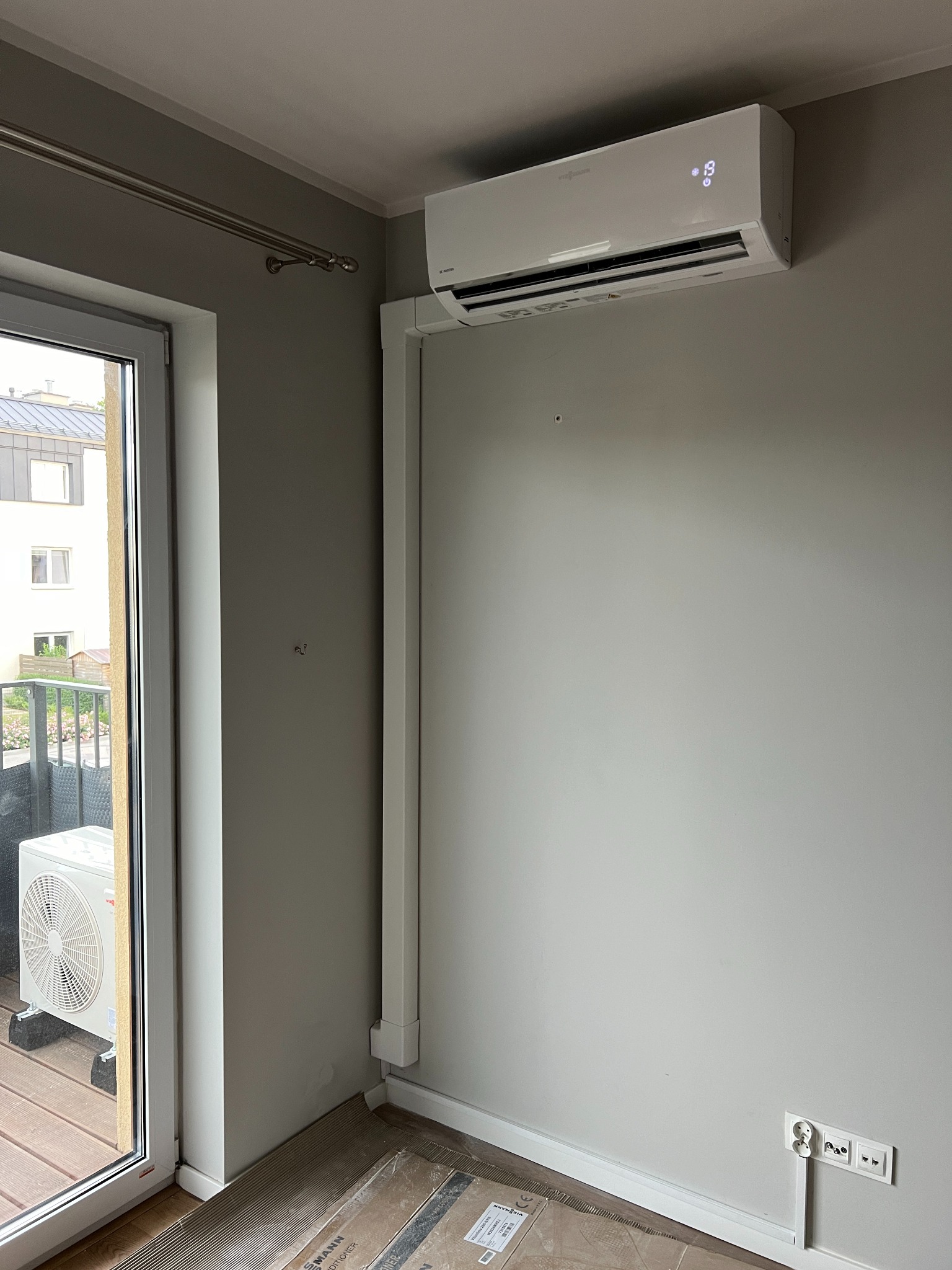 Biały klimatyzator marki Viessmann zamontowany na szarej ścianie w pokoju z balkonem; widoczna jednostka zewnętrzna na balkonie i zabezpieczona podłoga.