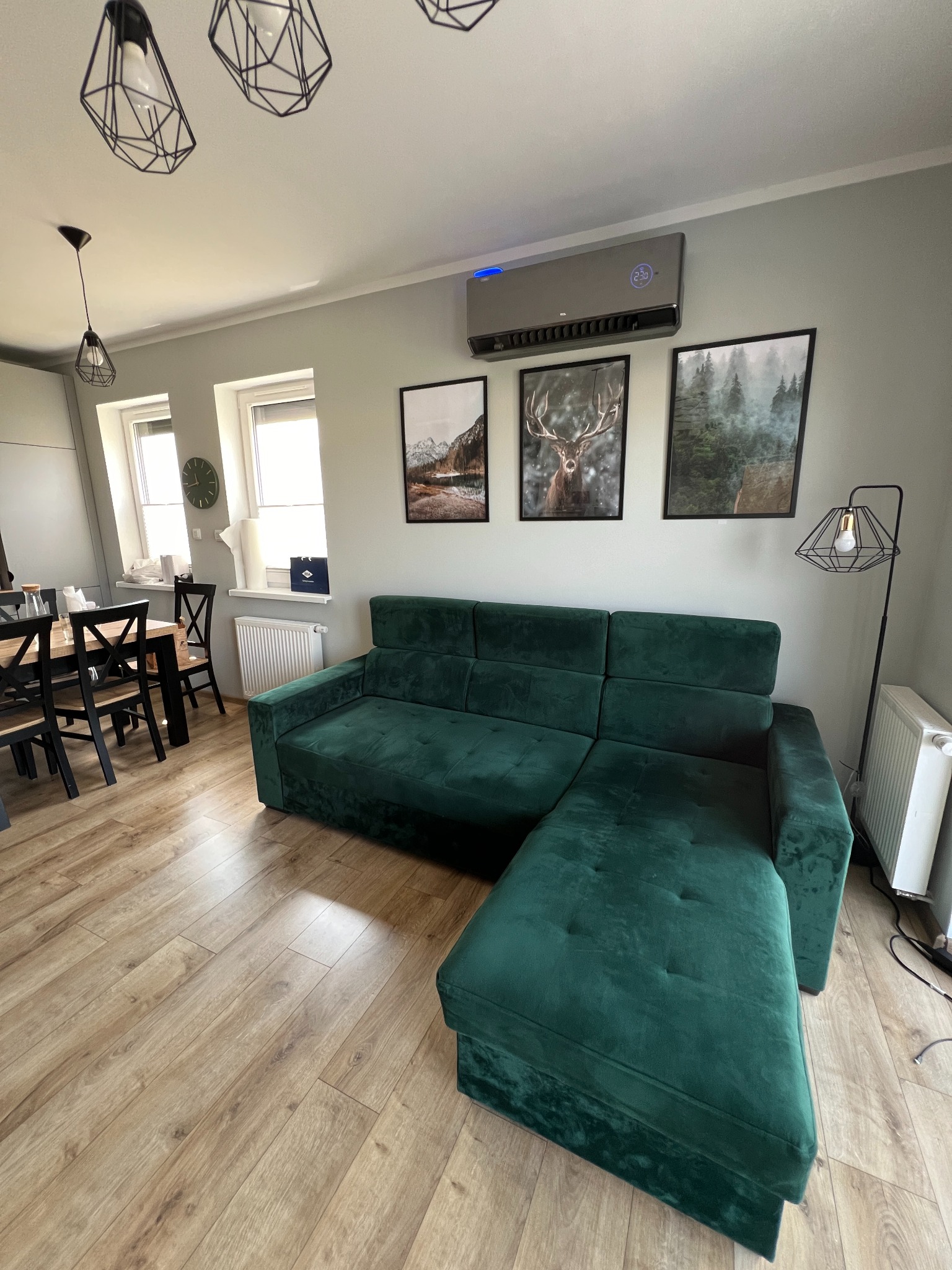 Szary klimatyzator ścienny z niebieskim podświetleniem, zamontowany nad zieloną, welurową sofą w nowoczesnym salonie z drewnianą podłogą i geometrycznymi lampami.
