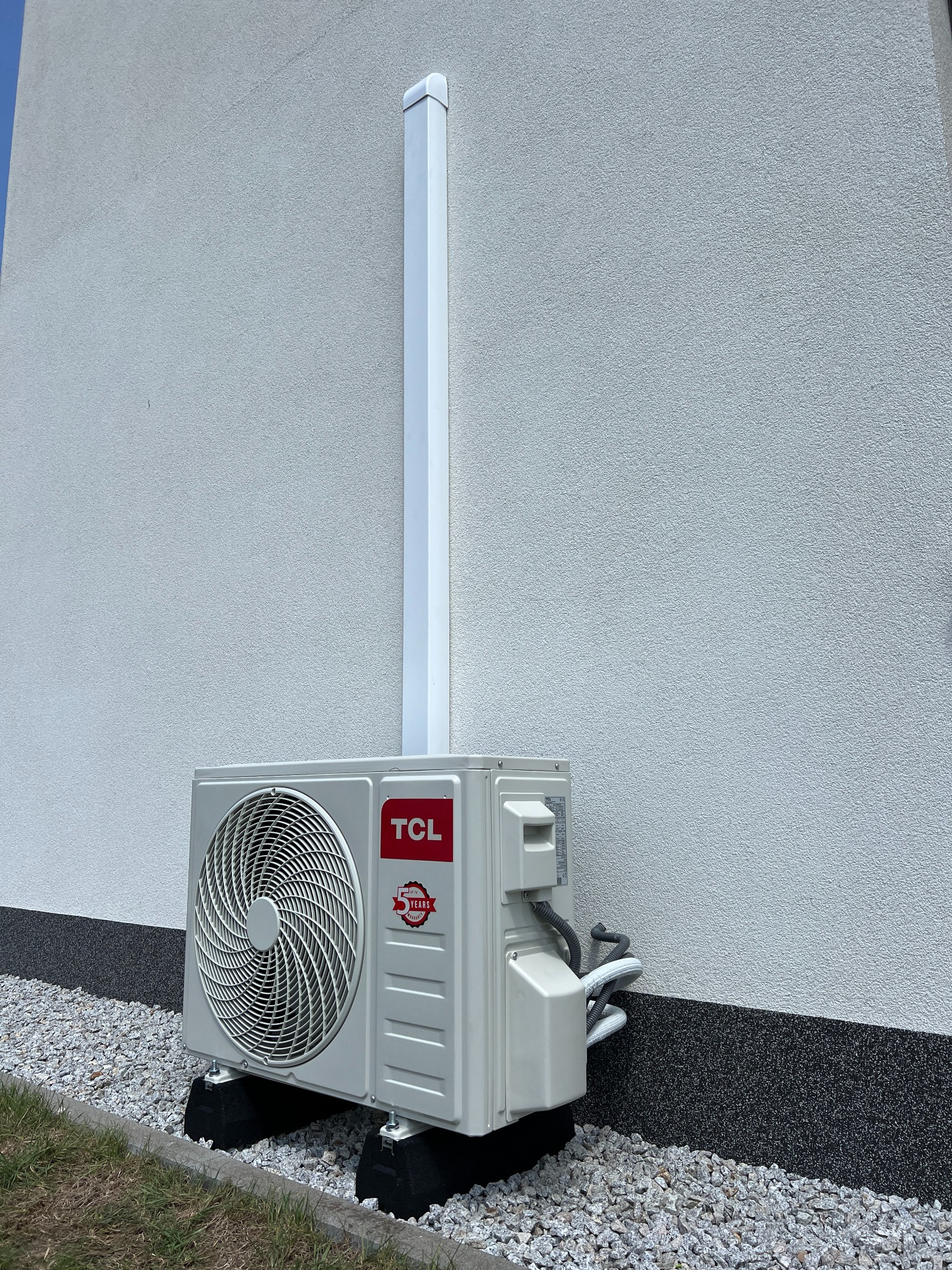 Zewnętrzna jednostka klimatyzacji, model TCL, zamontowana przy ścianie budynku z szarym tynkiem, na podłożu z drobnych kamieni. Widoczna biała rura odprowadzająca skropliny.