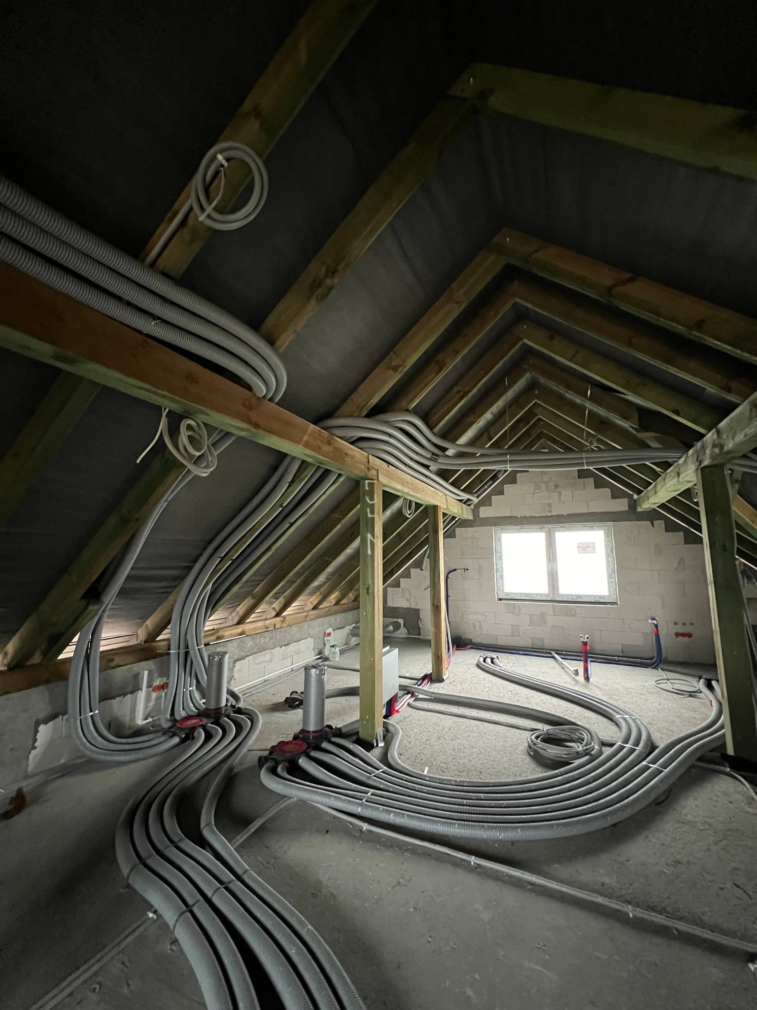 Rozprowadzenie szarych rur instalacyjnych w surowym stanie poddasza, widoczne drewniane elementy konstrukcyjne dachu i białe bloczki ścian, okno w tle.