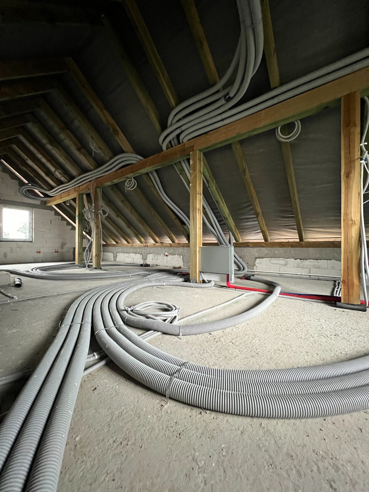 Szare, karbowane rury instalacyjne prowadzone pod drewnianą konstrukcją dachu na poddaszu w surowym stanie; widoczna skrzynka elektryczna na ścianie z niedokończonymi fragmentami instalacji.