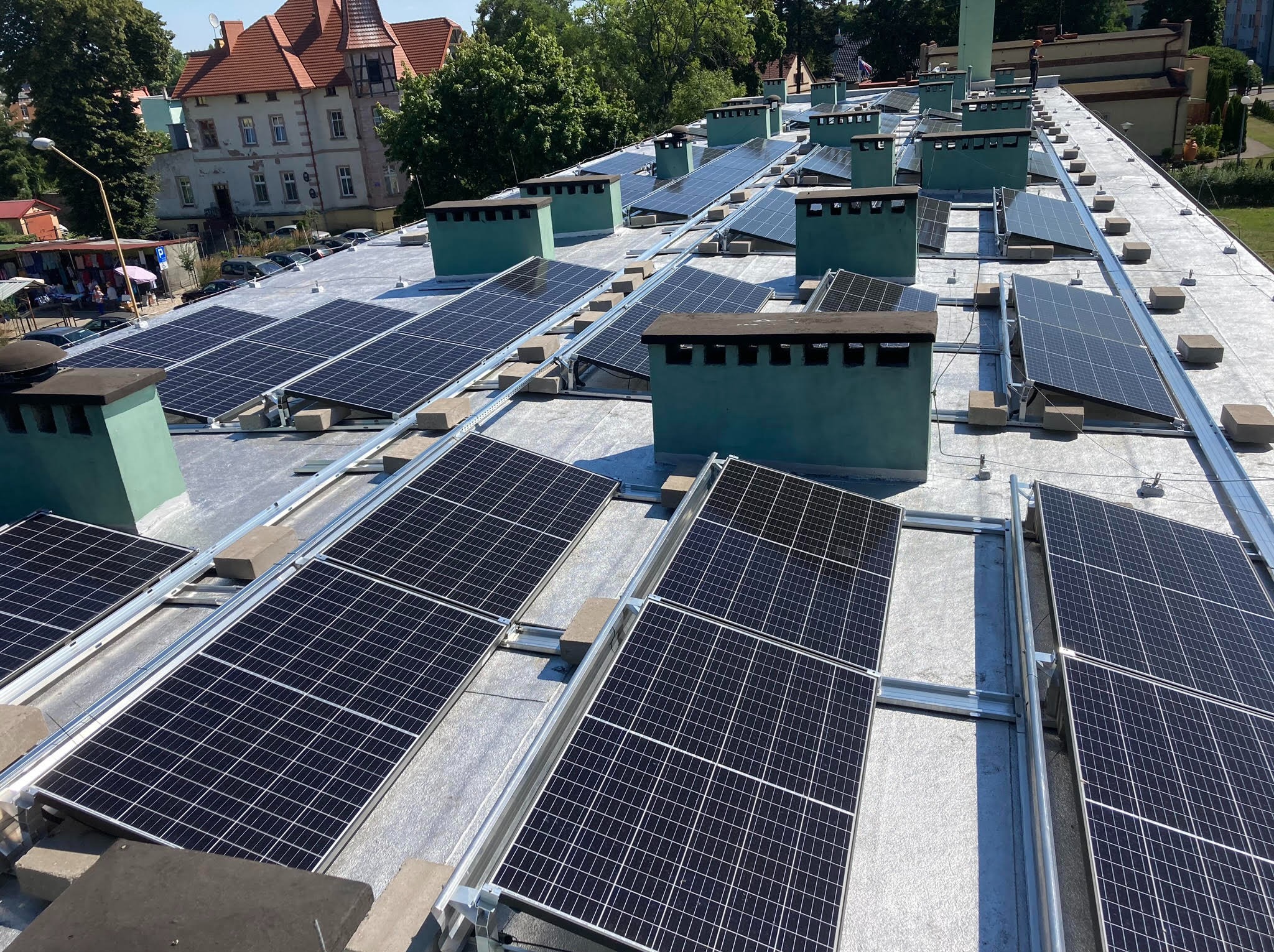 Instalacja paneli słonecznych na płaskim dachu budynku z zielonymi kominami wentylacyjnymi, widok z góry, w tle budynki miejskie.