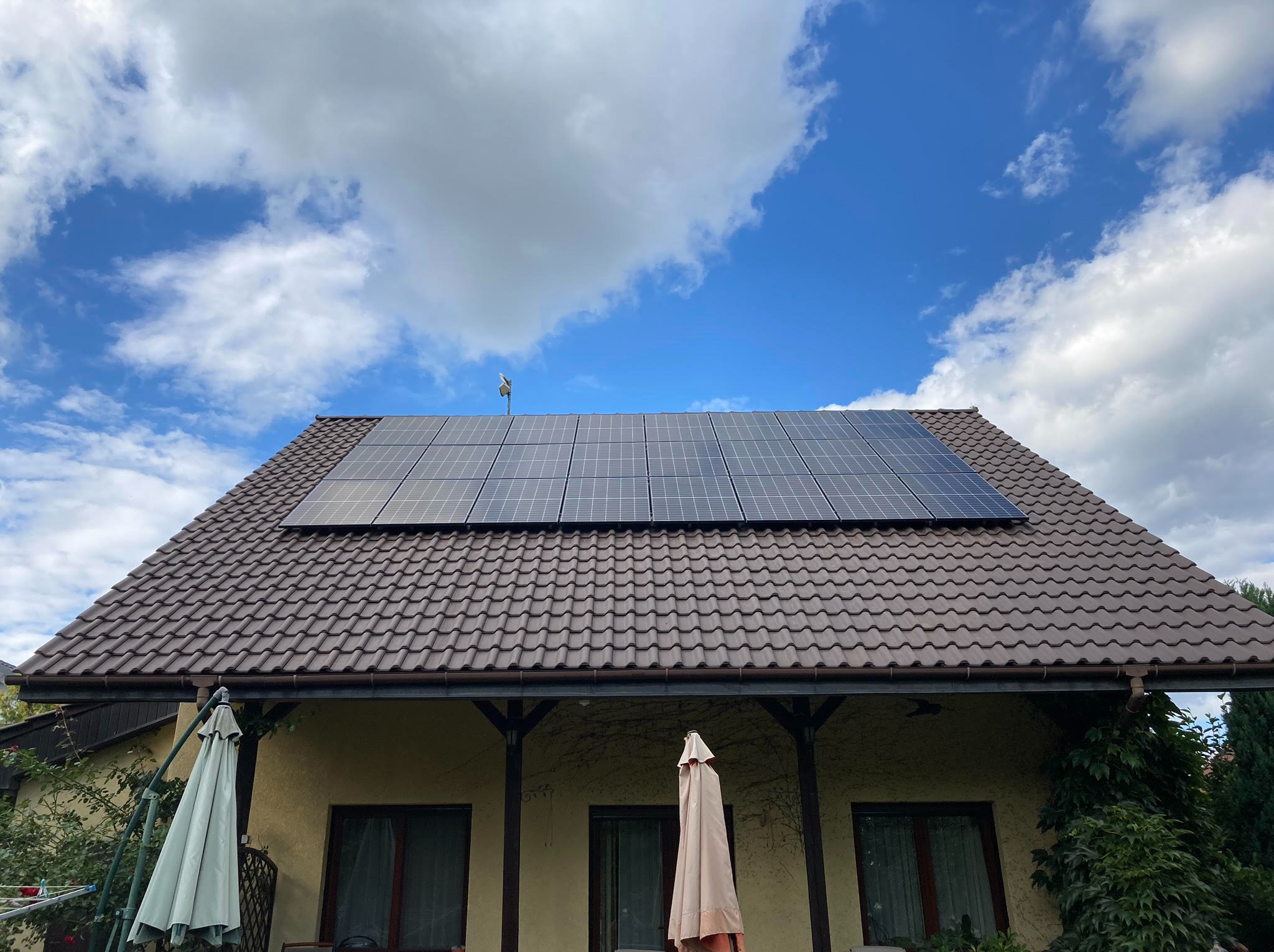 Panele fotowoltaiczne na brązowym dachu domu jednorodzinnego, błękitne niebo z białymi chmurami w tle, widoczna antena na dachu.