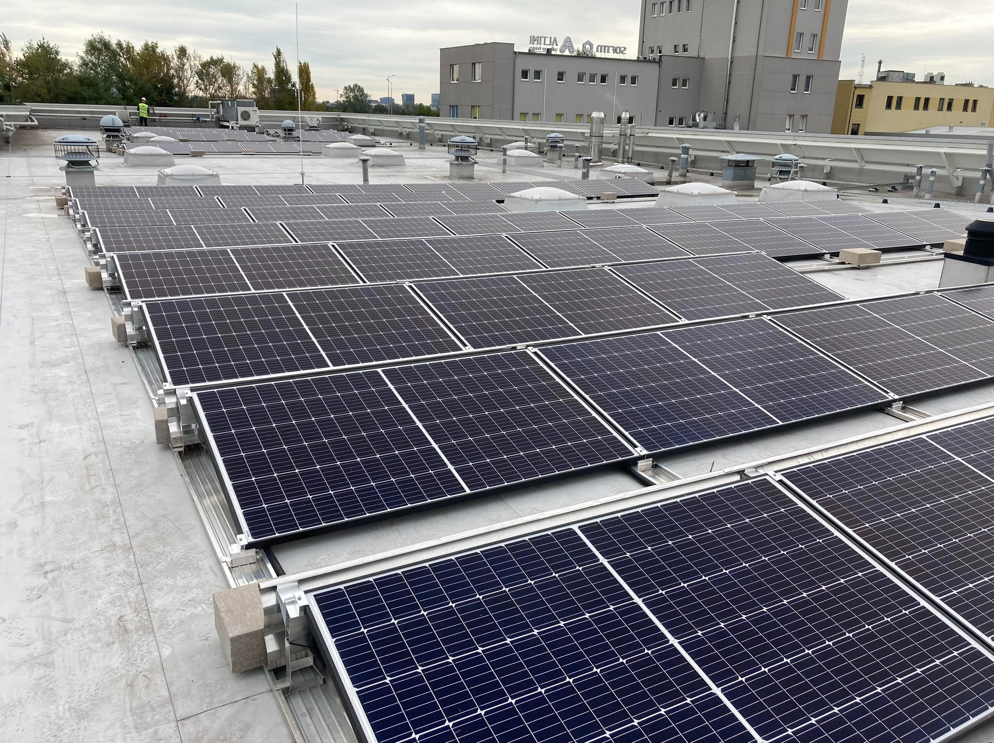 Rząd paneli słonecznych na dachu budynku przemysłowego, widoczny pracownik w odblaskowej kamizelce, szare niebo.