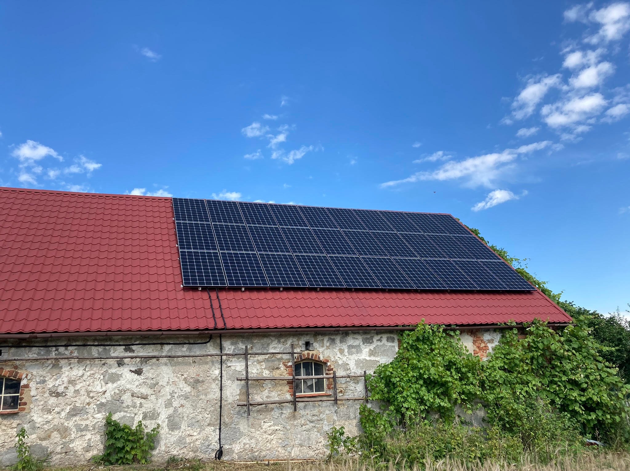Panele słoneczne na czerwonym dachu starego, kamiennego budynku gospodarczego w słoneczny dzień.