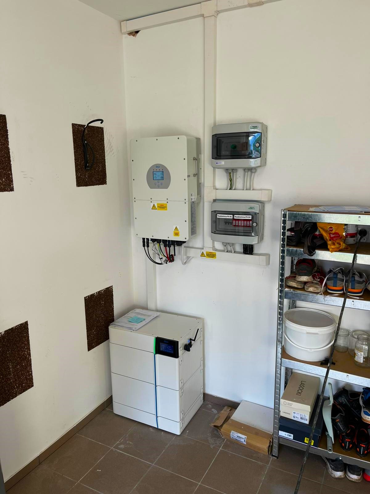 Zamontowany biały falownik fotowoltaiczny z podłączonymi przewodami, skrzynki zabezpieczeń AC i DC oraz modułowy magazyn energii z zielonym akcentem, ustawione w pomieszczeniu gospodarczym.