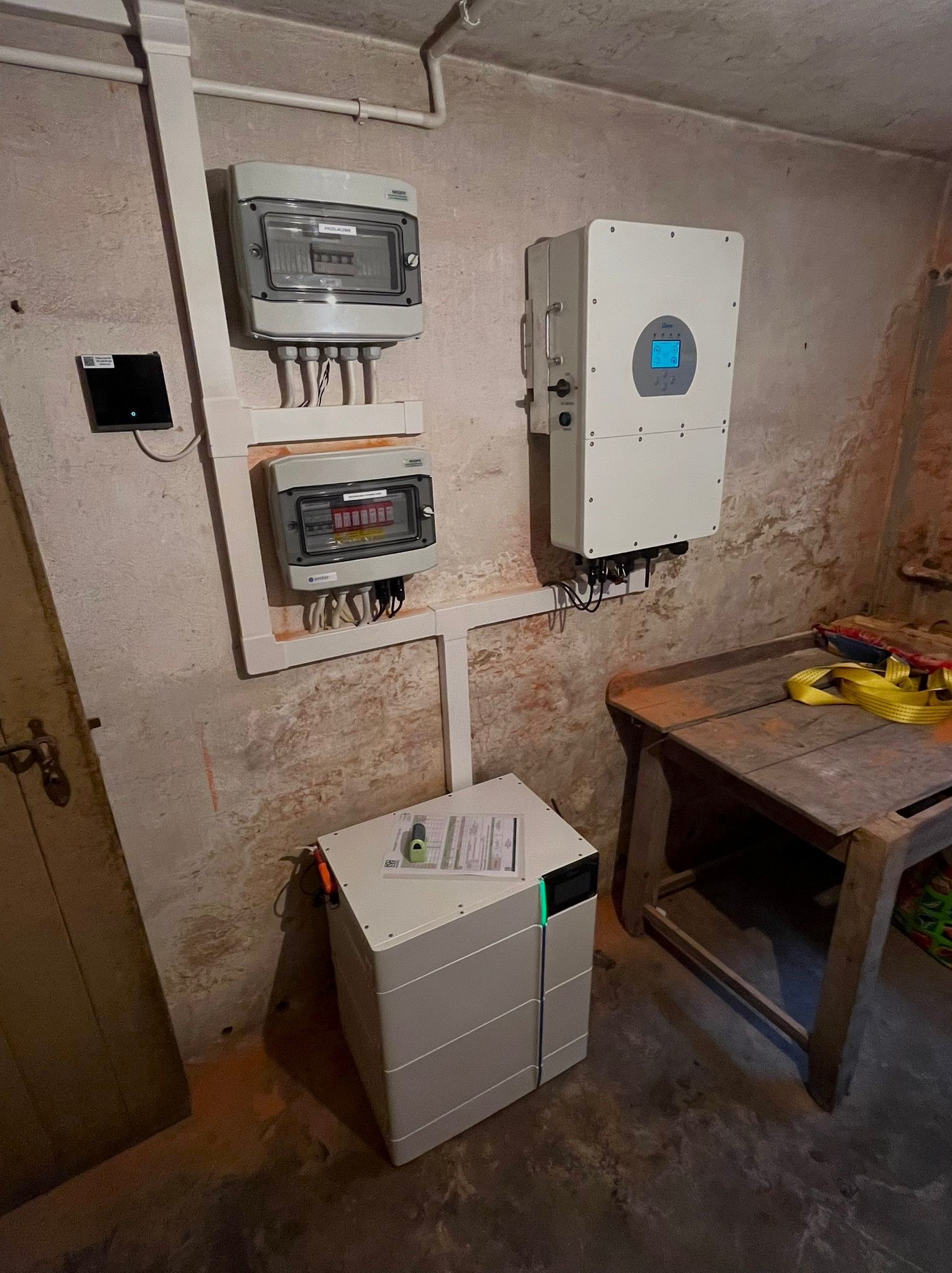 Wnętrze z zamontowanym inwerterem, dwoma rozdzielnicami elektrycznymi i akumulatorem energii na tle surowej ściany z widoczną instalacją elektryczną w białych rurkach.