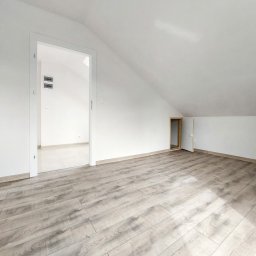 Panarent sp. z o.o. - Poddasze po remoncie: jasne panele podłogowe imitujące drewno, białe ściany i skośny sufit z dwoma otworami drzwiowymi, jeden prowadzący do pomieszczenia z białymi płytkami podłogowymi.