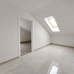 Panarent sp. z o.o. - Jasne poddasze z białymi ścianami, ceramiczną podłogą w odcieniach beżu i oknem dachowym z drewnianą ramą, widok na sąsiednie pomieszczenie z panelami podłogowymi.