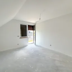 Pusty pokój na poddaszu z oknem i surową, betonową podłogą przygotowaną pod wylewkę. Ściany pomalowane na biało, widoczne gniazdka elektryczne. Na suficie wisi żarówka na kablu.