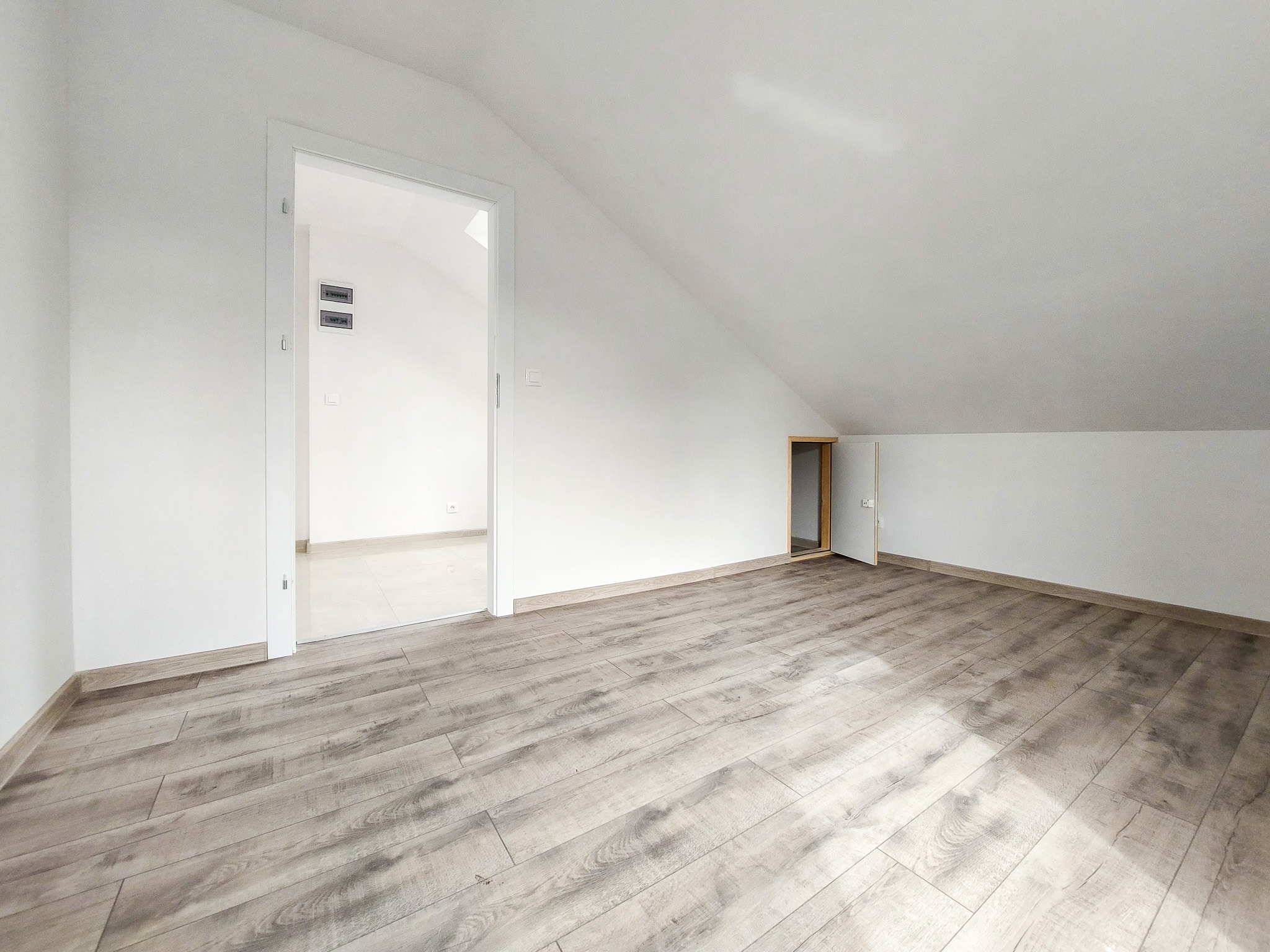 Poddasze po remoncie: jasne panele podłogowe imitujące drewno, białe ściany i skośny sufit z dwoma otworami drzwiowymi, jeden prowadzący do pomieszczenia z białymi płytkami podłogowymi.
