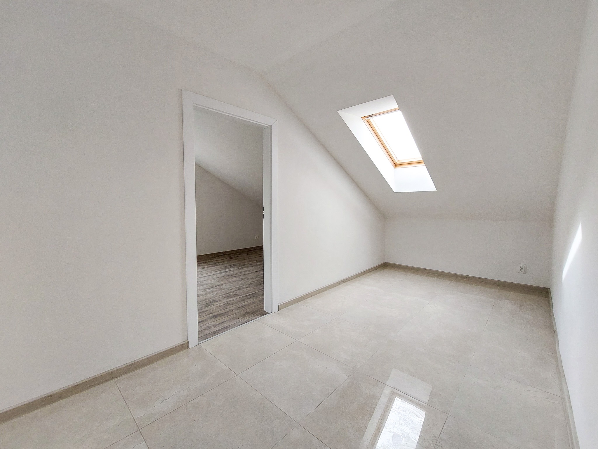 Jasne poddasze z białymi ścianami, ceramiczną podłogą w odcieniach beżu i oknem dachowym z drewnianą ramą, widok na sąsiednie pomieszczenie z panelami podłogowymi.