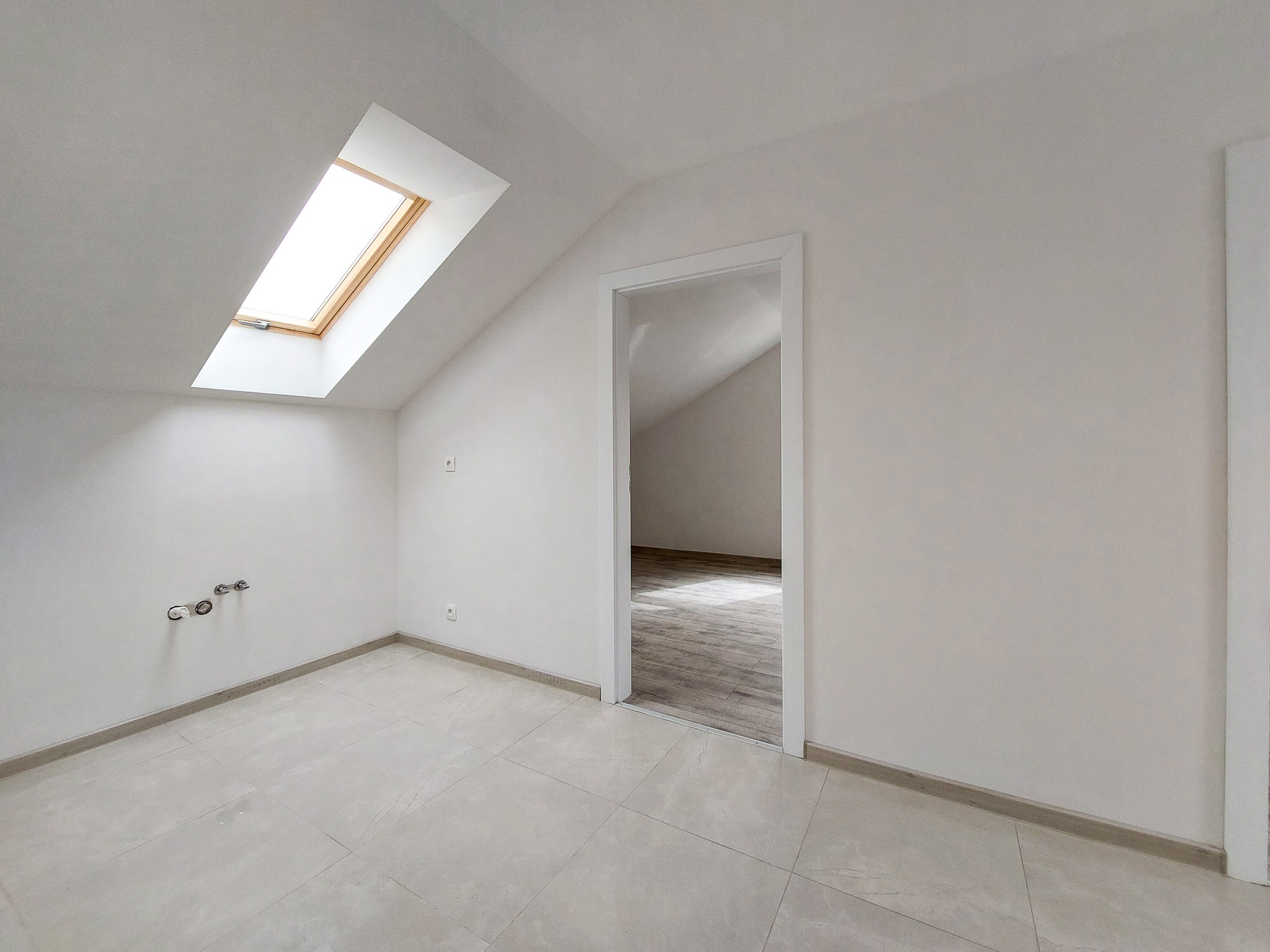 Poddasze po remoncie z oknem dachowym, świeżo pomalowane na biało, z jasnymi płytkami na podłodze i przygotowanymi przyłączami hydraulicznymi, widok na sąsiednie pomieszczenie z parkietem.