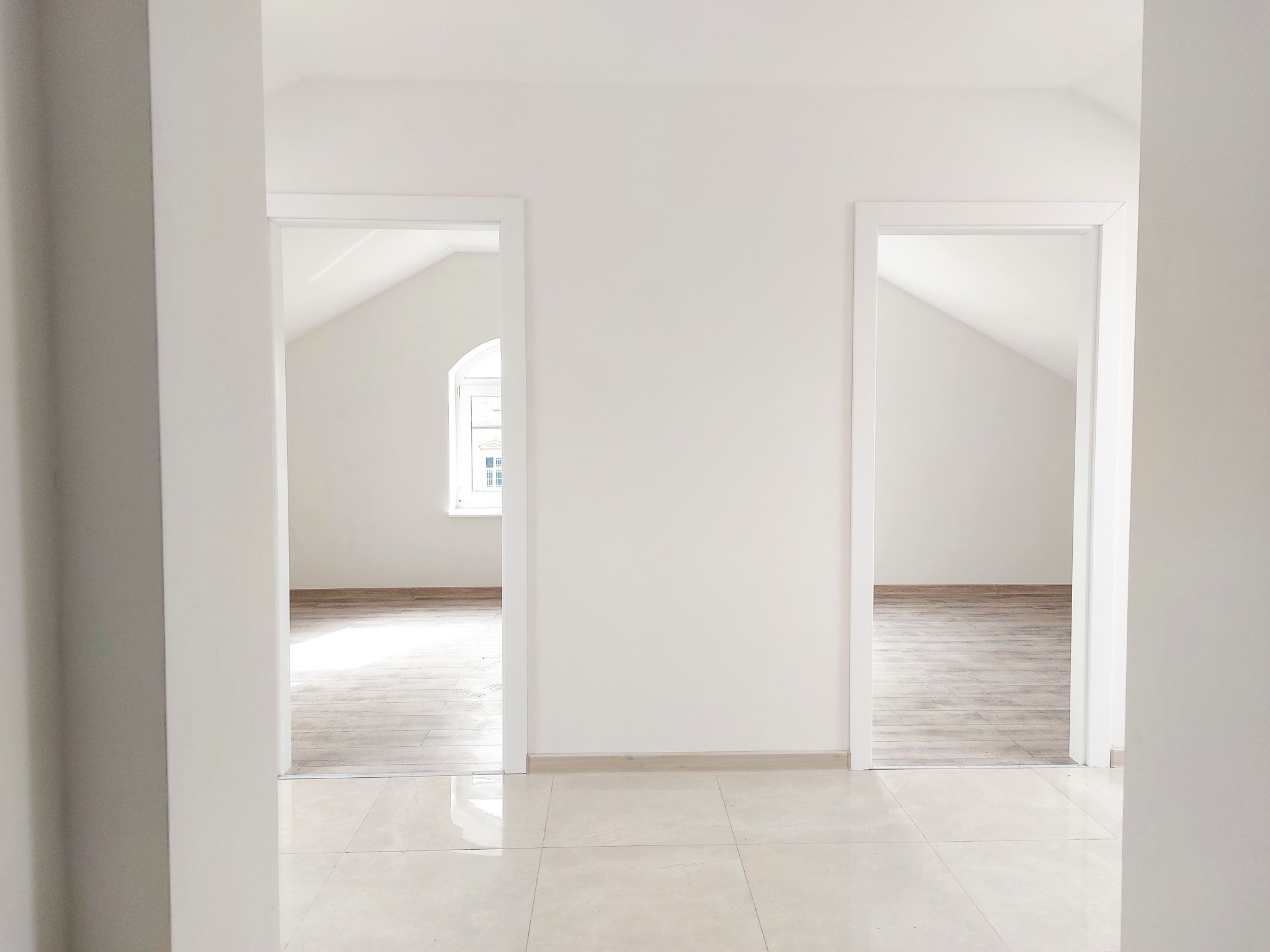 Jasne, minimalistyczne wnętrze z białymi ścianami, dwoma otwartymi przejściami z białymi ościeżnicami, kremową podłogą z płytek i widocznym fragmentem podłogi z paneli w pokojach w głębi.