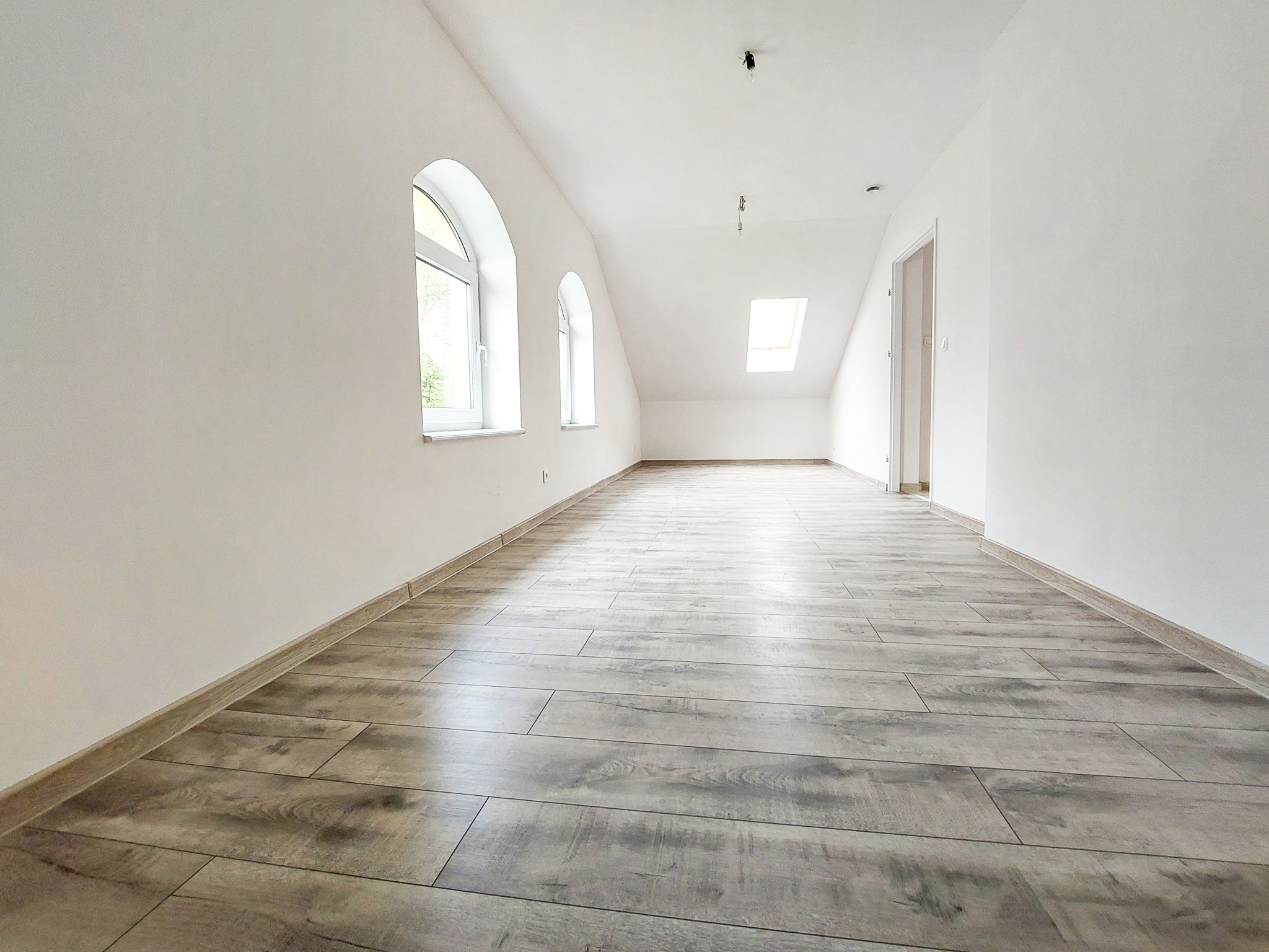 Poddasze po remoncie: jasne ściany, szare panele podłogowe imitujące drewno, dwa okna łukowe i okno dachowe.