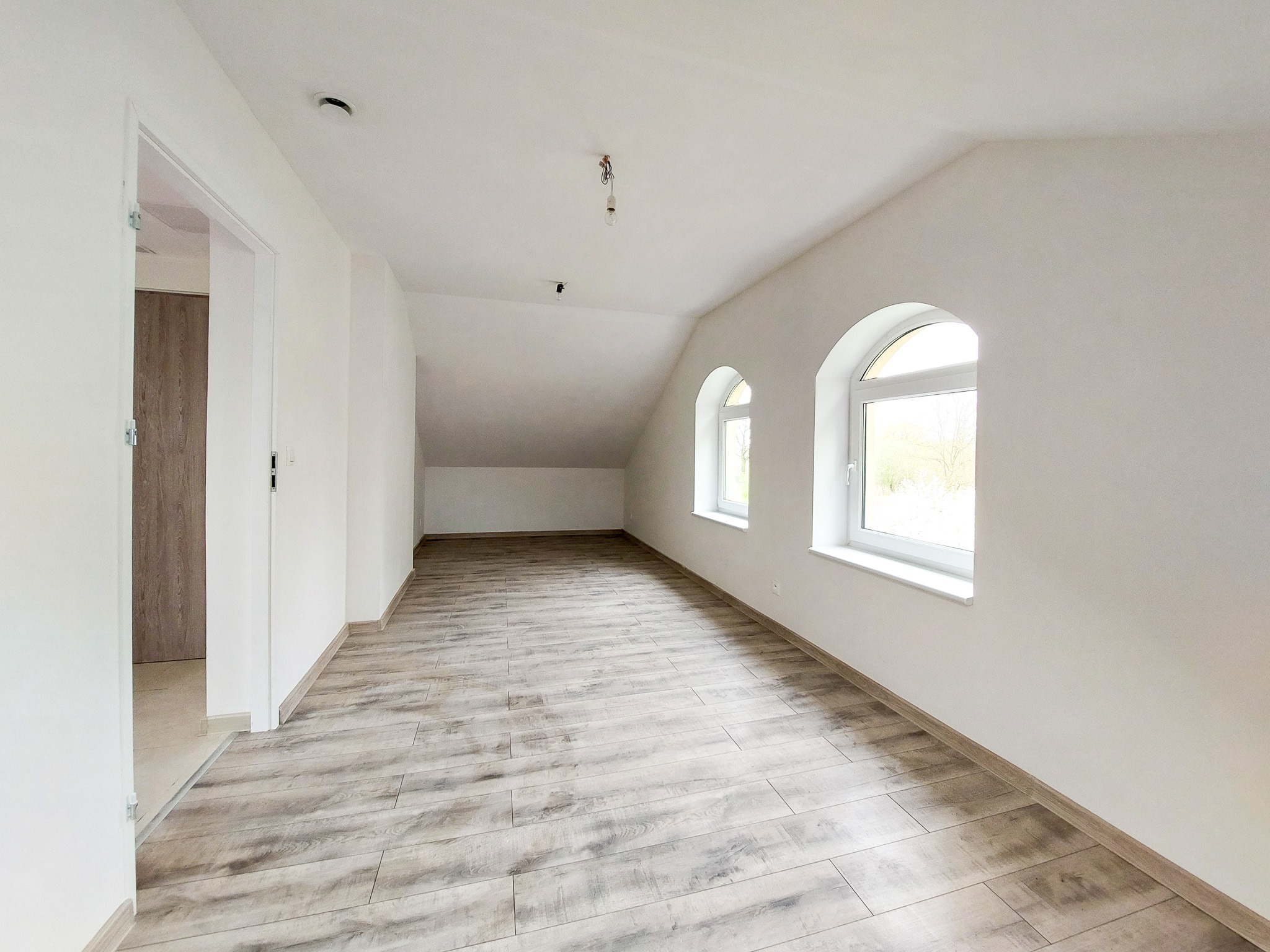 Poddasze po remoncie z nową podłogą imitującą drewno, białe ściany i dwa łukowe okna w jasnych ramach, widoczny fragment instalacji elektrycznej sufitowej.