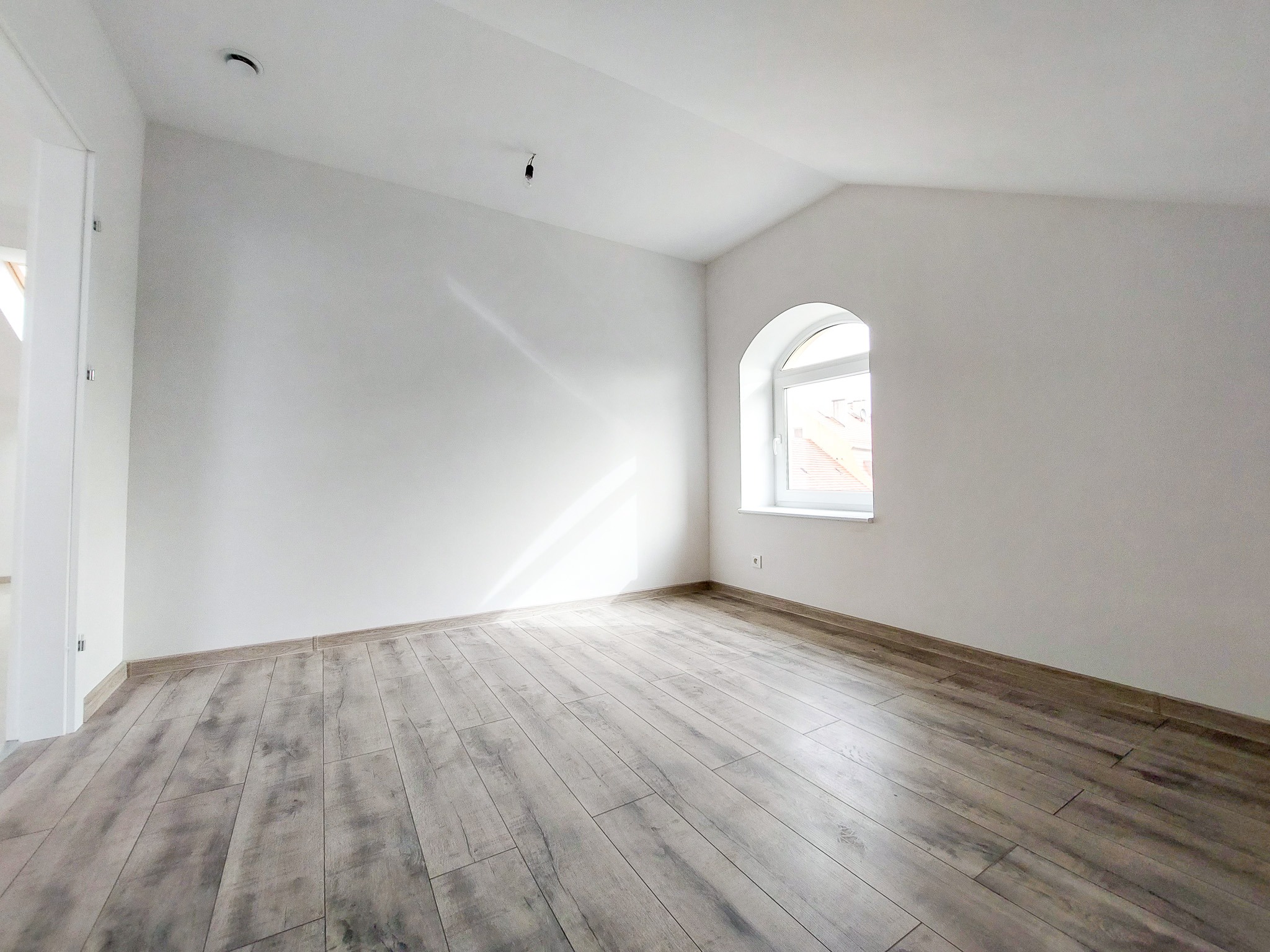 Jasne, puste pomieszczenie na poddaszu z ułożoną podłogą z paneli imitujących drewno, białymi ścianami, oknem łukowym i przygotowanym punktem oświetleniowym na suficie.
