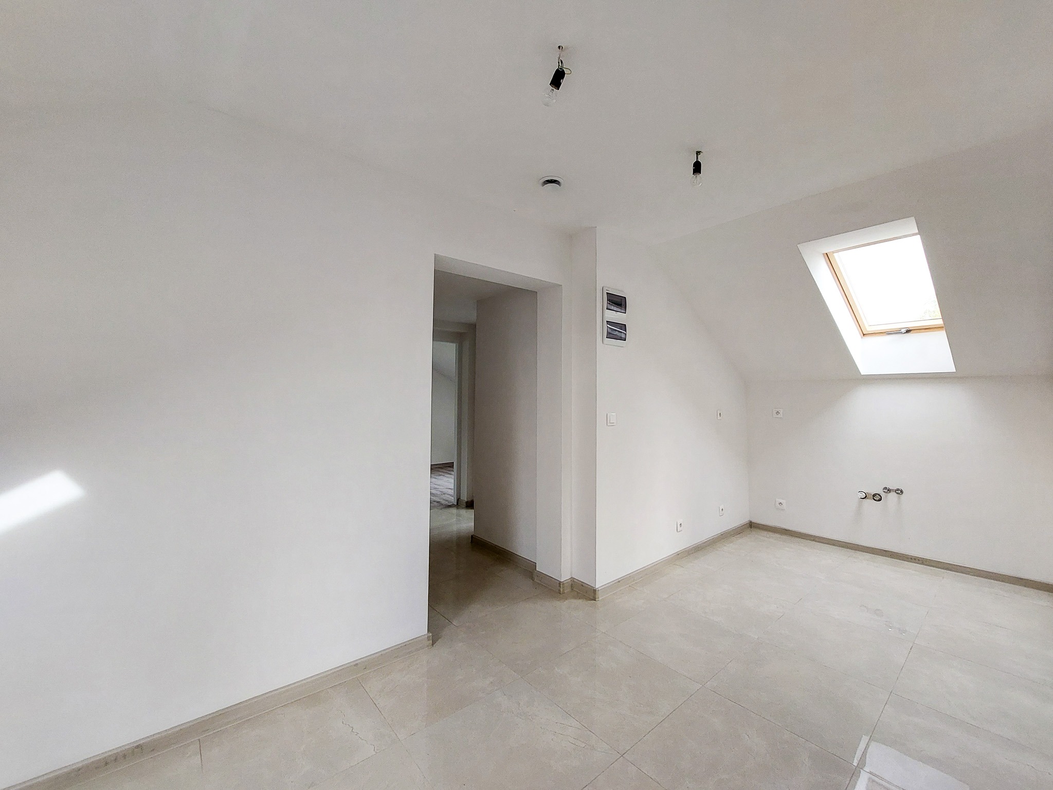 Poddasze po remoncie: świeżo pomalowane białe ściany, beżowe płytki podłogowe, okno dachowe i instalacja elektryczna bez opraw oświetleniowych.