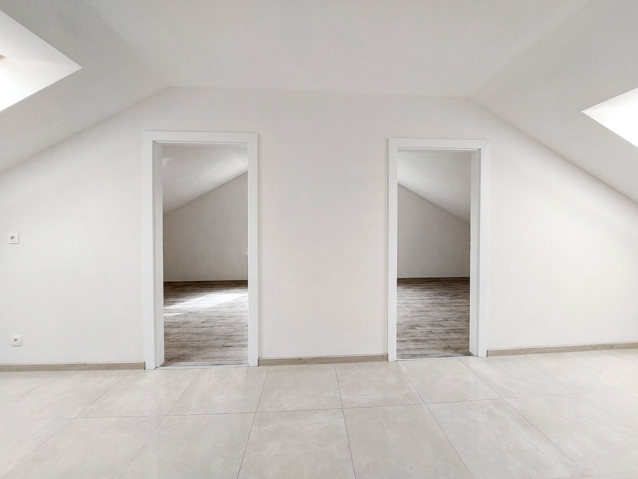 Poddasze po remoncie z dwoma wejściami do pokojów, białe ściany, jasna, kwadratowa podłoga z płytek, widoczne okna dachowe.