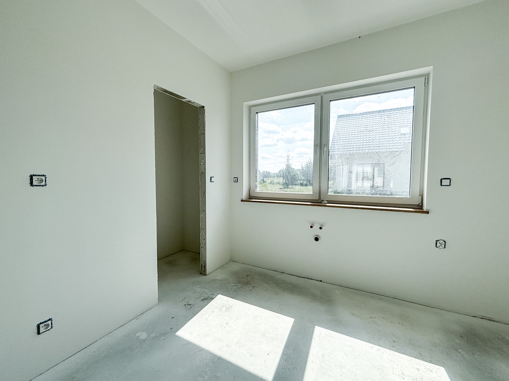 Surowe wnętrze pokoju w trakcie remontu: białe ściany, widoczne puszki elektryczne, otwór drzwiowy bez ościeżnicy, betonowa podłoga z plamami, okno z widokiem na dom i zieleń.