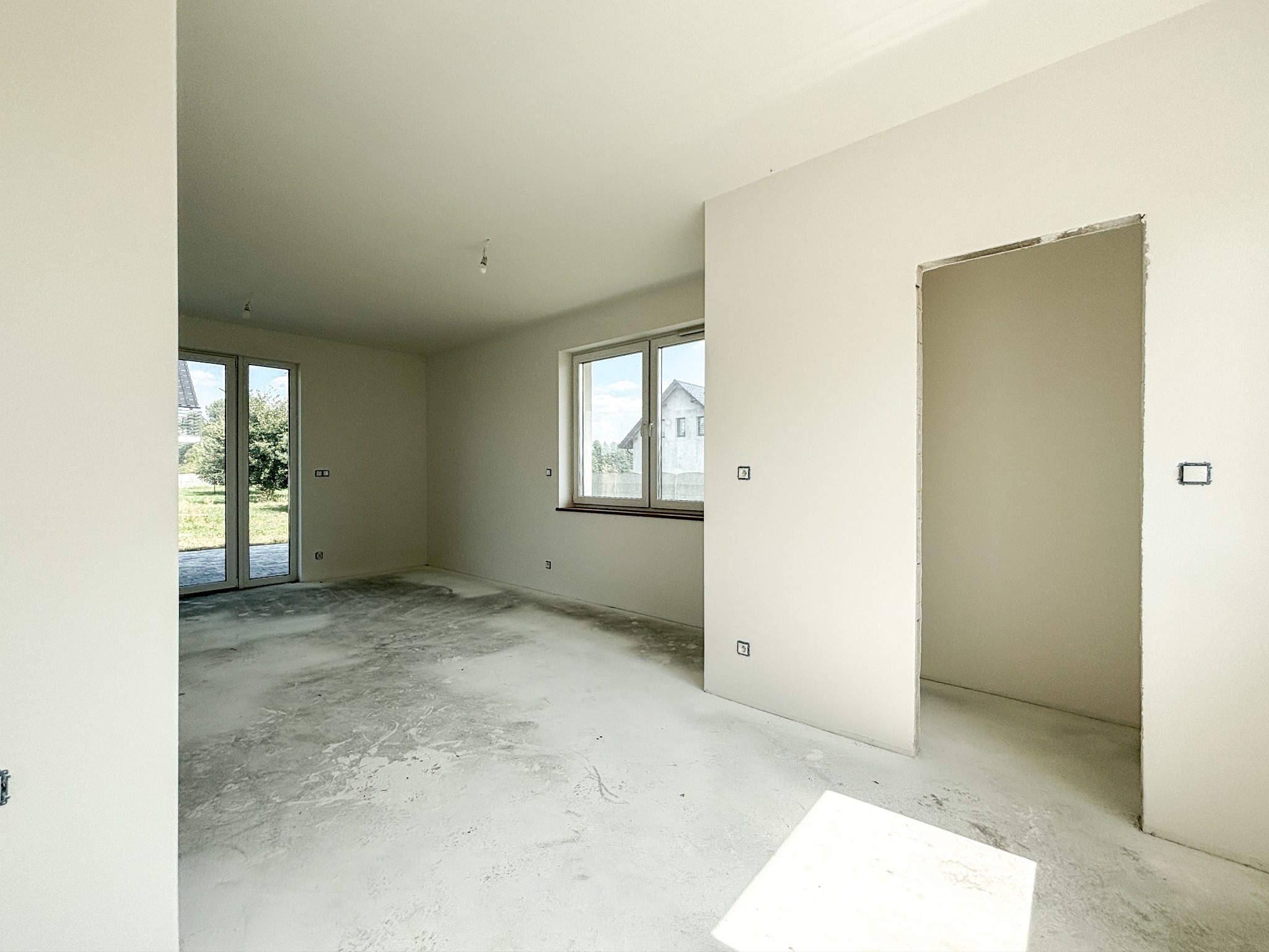 Surowe wnętrze w stanie deweloperskim z gładkimi, pomalowanymi na biało ścianami, surową betonową podłogą, oknami z widokiem na zieleń i niezamaskowanymi puszkami elektrycznymi.