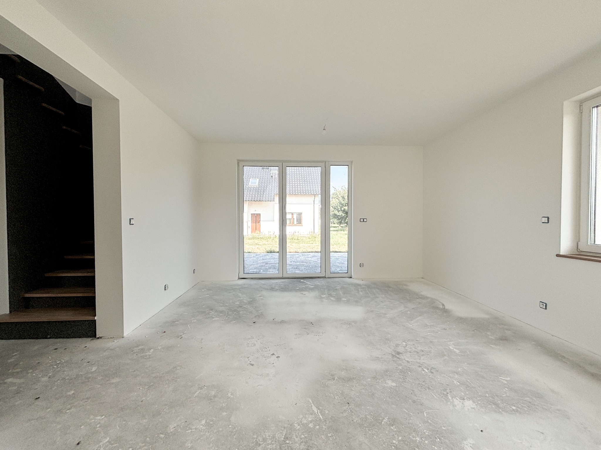 Wnętrze surowego stanu z białymi ścianami, betonową posadzką i widocznymi schodami z czarną balustradą, przez okno widoczny dom.