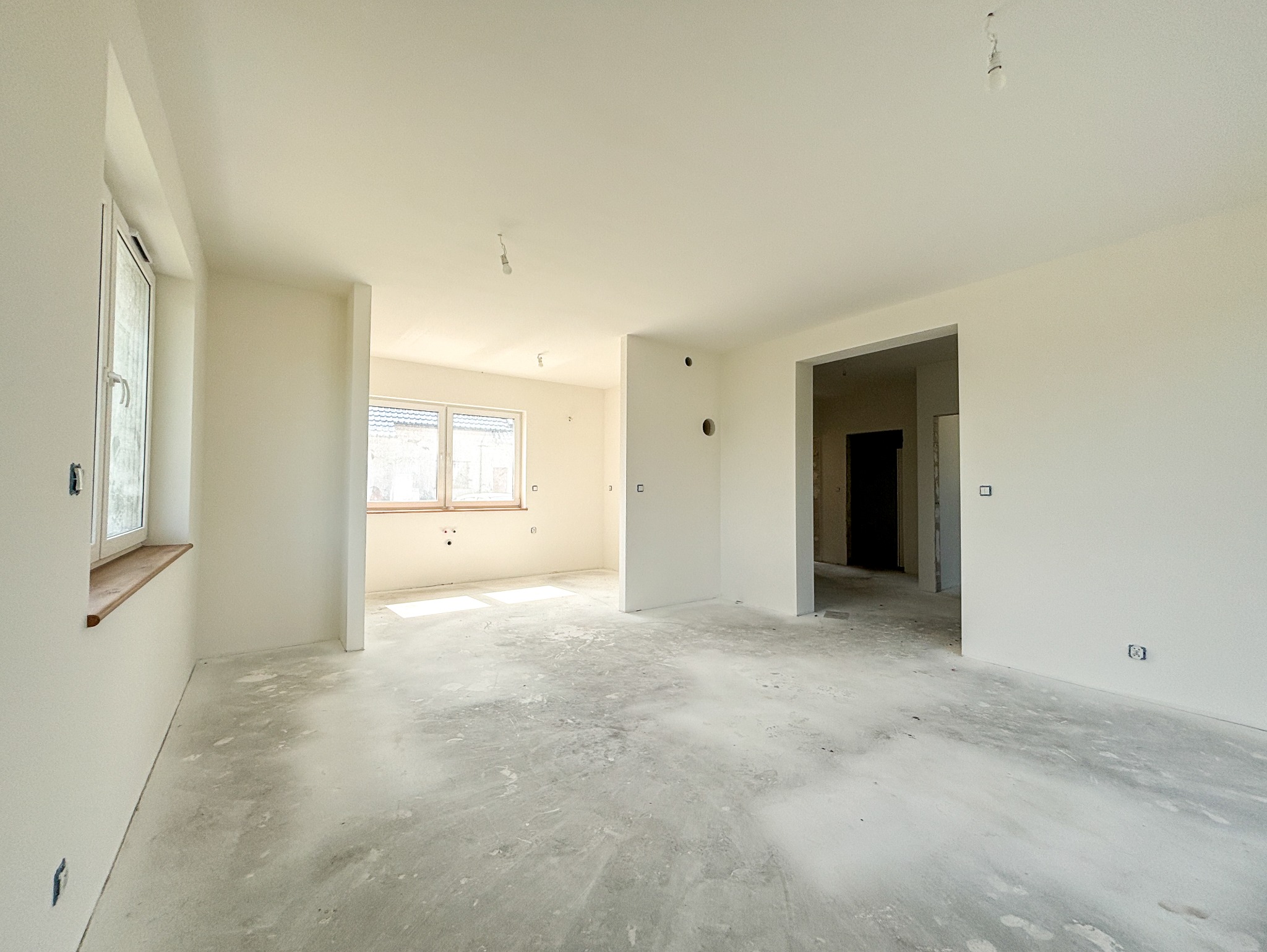 Puste, jasne wnętrze mieszkania w trakcie prac wykończeniowych, widoczne ślady po pracach na podłodze, świeżo pomalowane ściany, brak mebli, surowy stan przed urządzeniem.