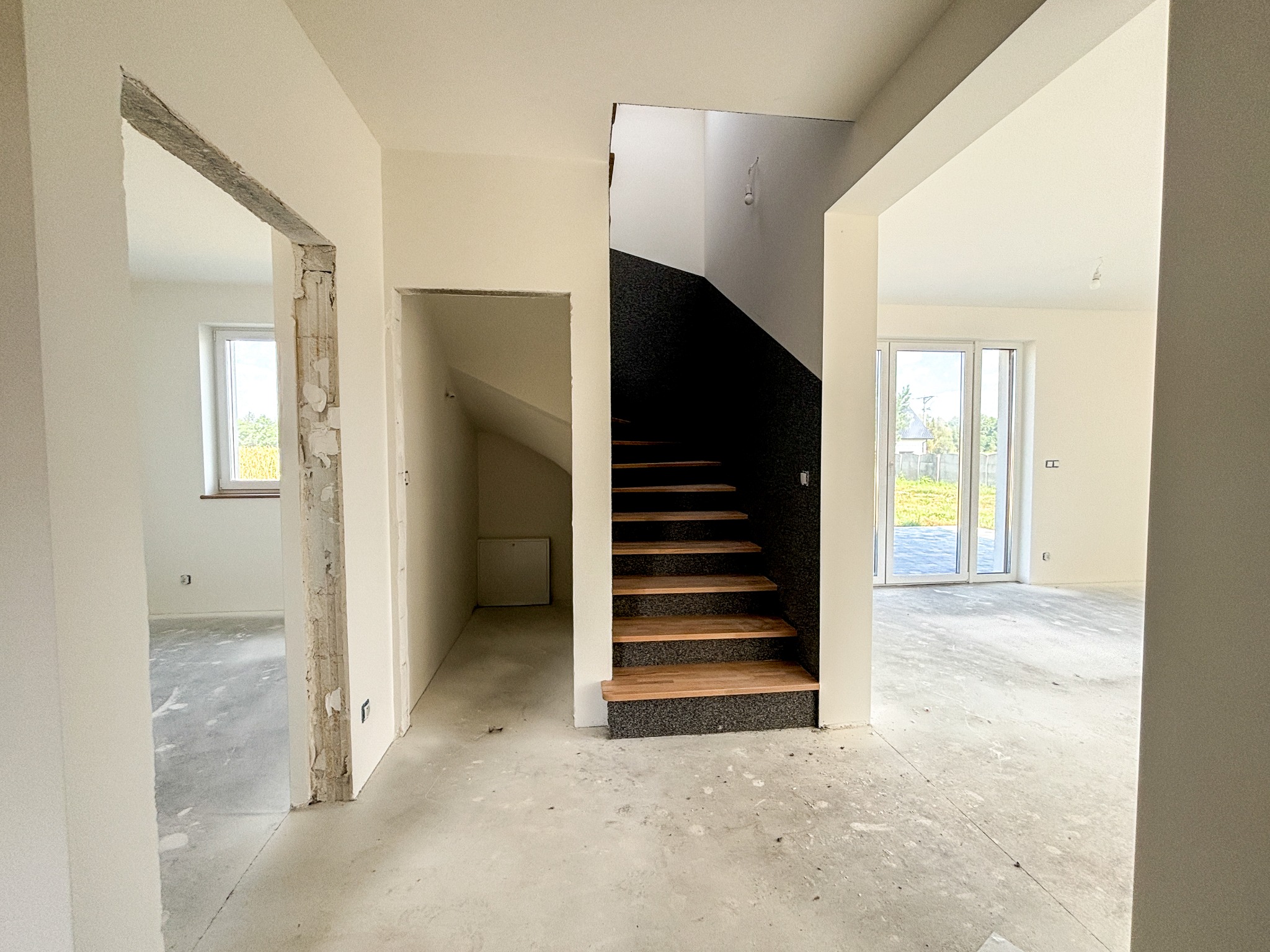 Wnętrze domu w trakcie prac wykończeniowych: widok na schody z drewnianymi stopniami i czarną poręczą, surowa podłoga, otwarte przestrzenie i widoczne okno.